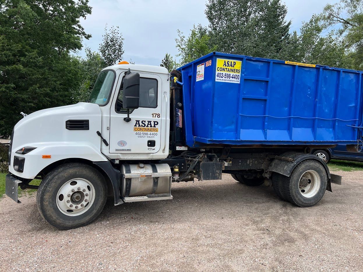 A white dump truck with a blue dumpster on the back is parked in a gravel lot.