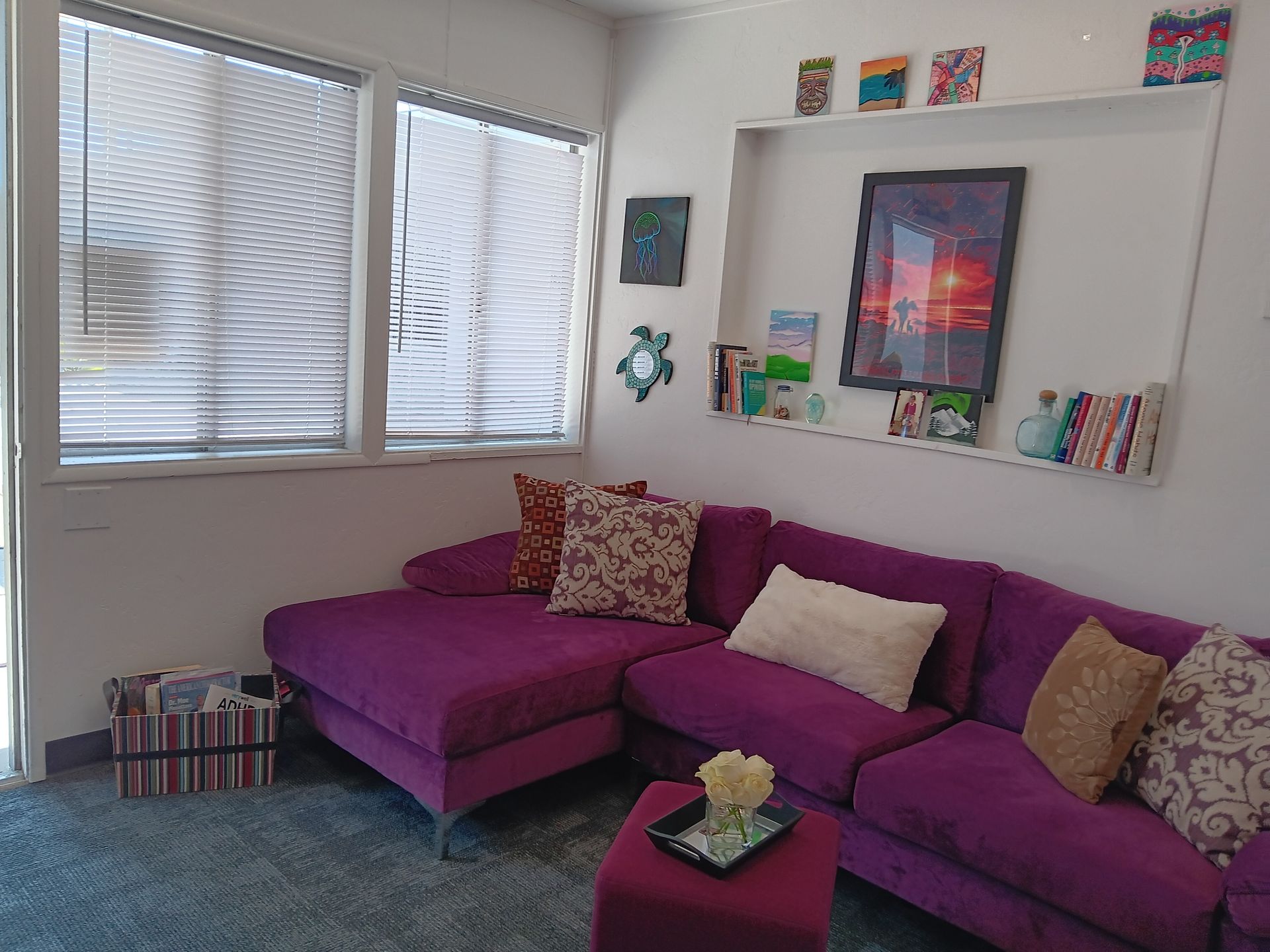 Cozy living room with purple sectional sofa, windows, built-in shelves with art, and decorative pillows.