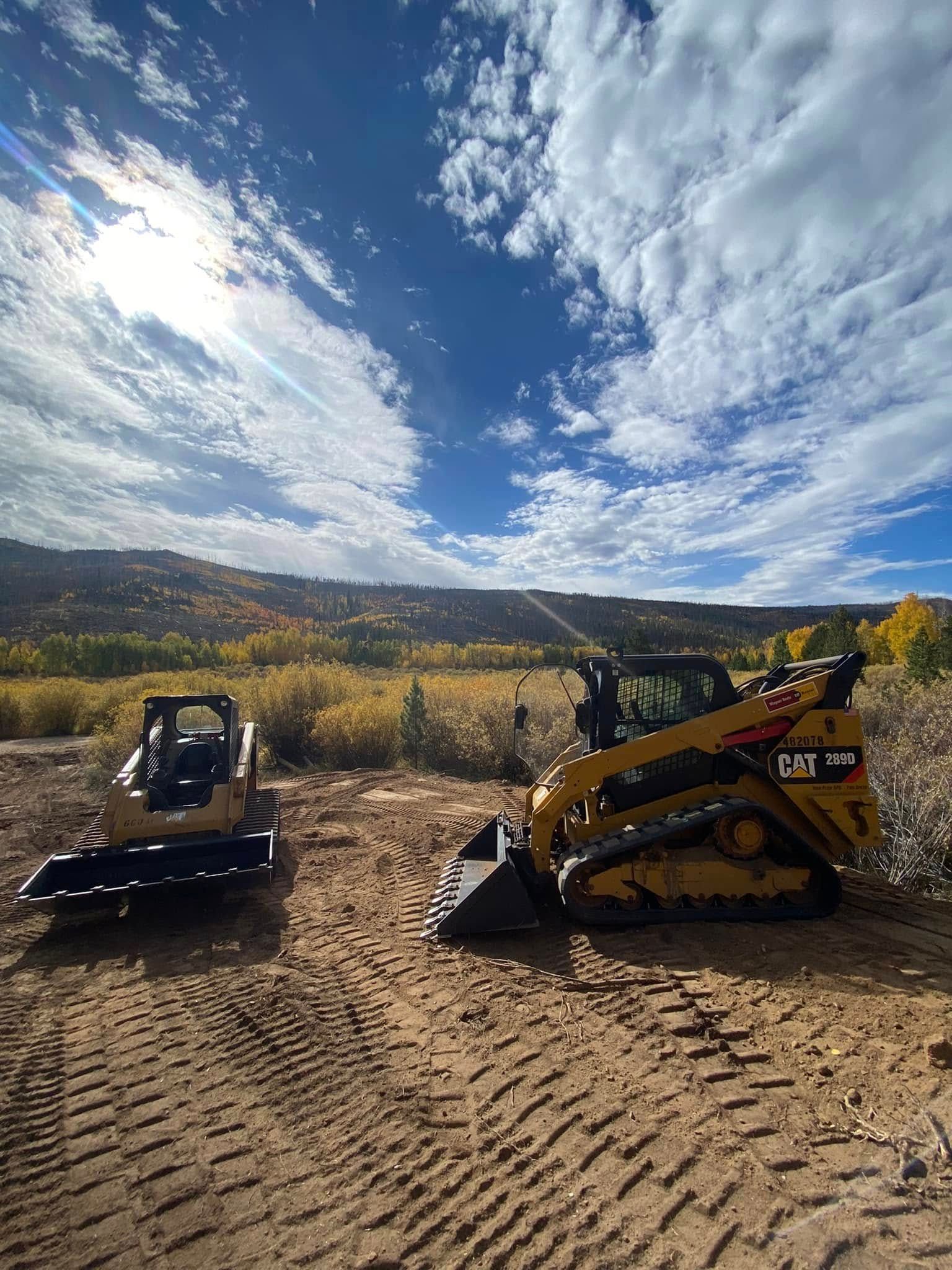 Two small excavators on a dirt lot under a bright, cloudy sky with mountains in the background