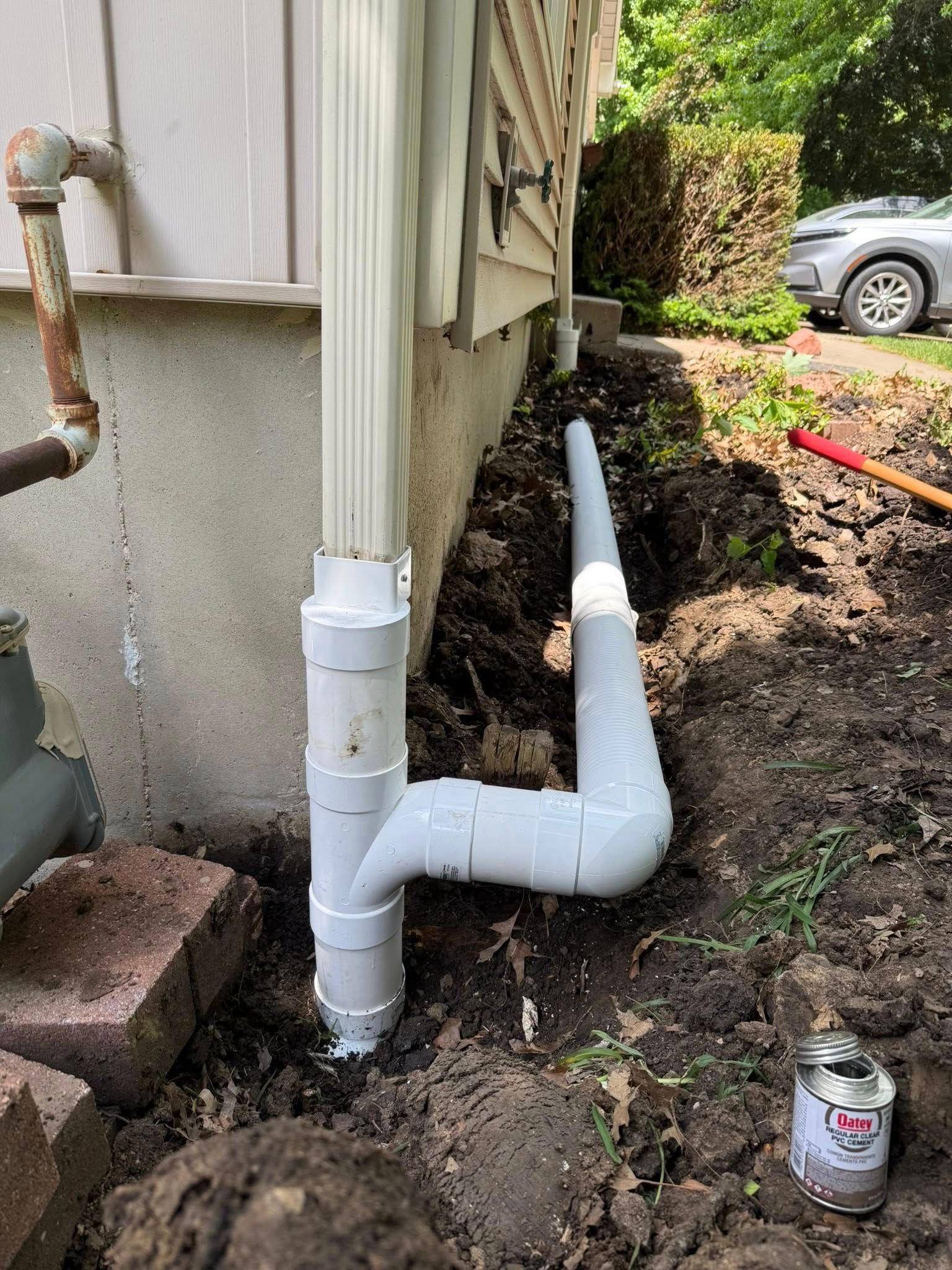 White PVC drainage pipes beside a house foundation in a dirt trench, with a soda can nearby.