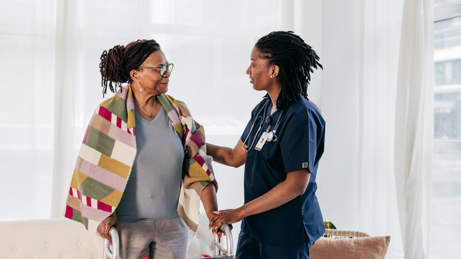 Caregiver in blue scrubs helps an older woman in a light blue sweater stand up from a sofa in a home.