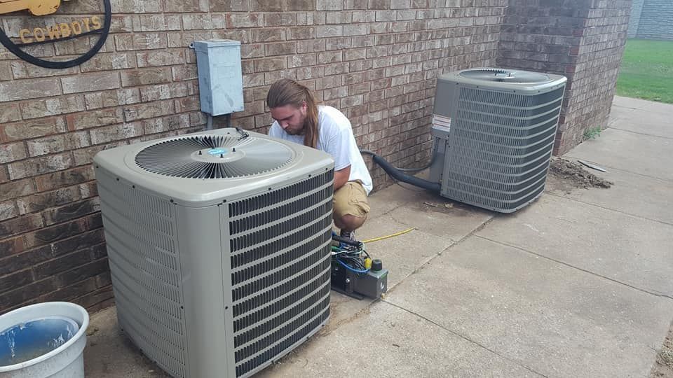 Person working on an air conditioning unit outside on concrete next to a brick building.