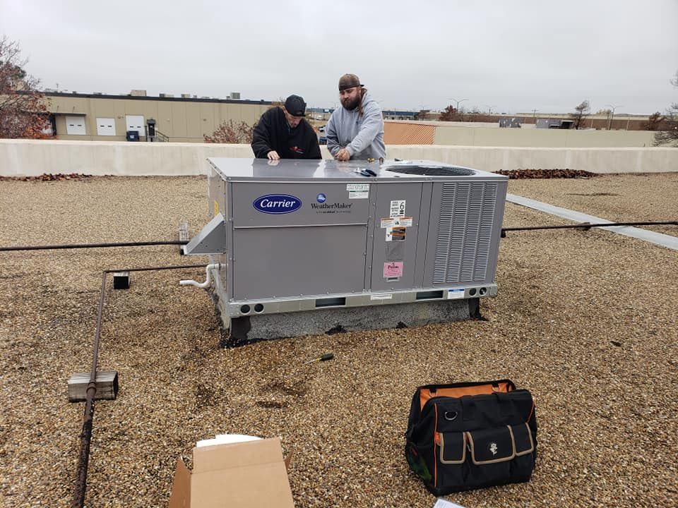 Two workers installing a Carrier rooftop HVAC unit on a gravel-covered flat roof.