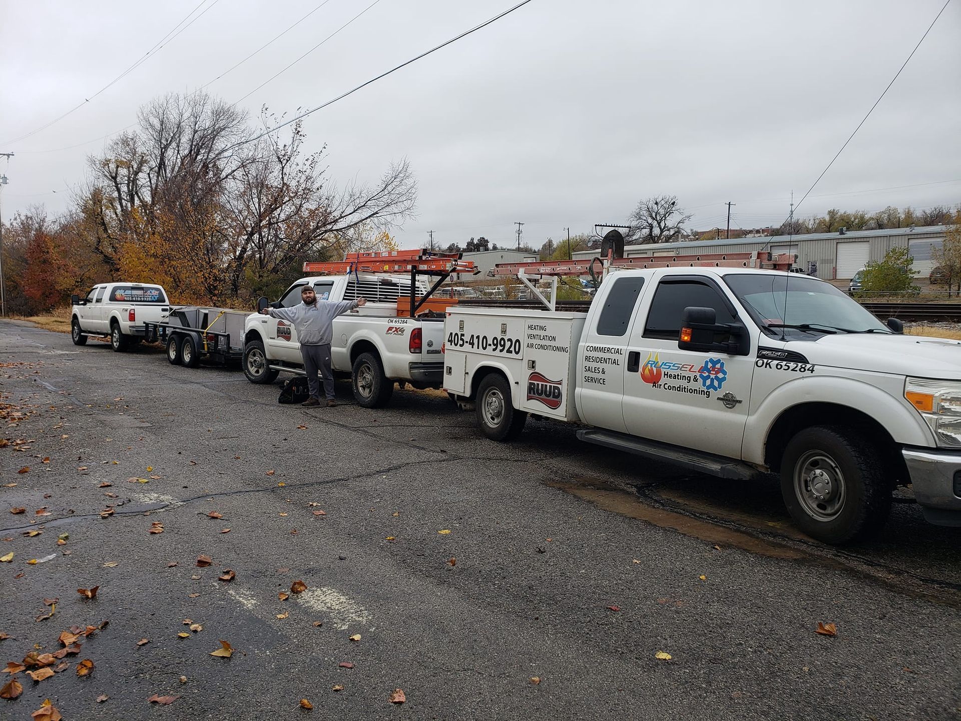 Three white work trucks with a trailer parked on a road. A person stands between two trucks. Overcast sky.