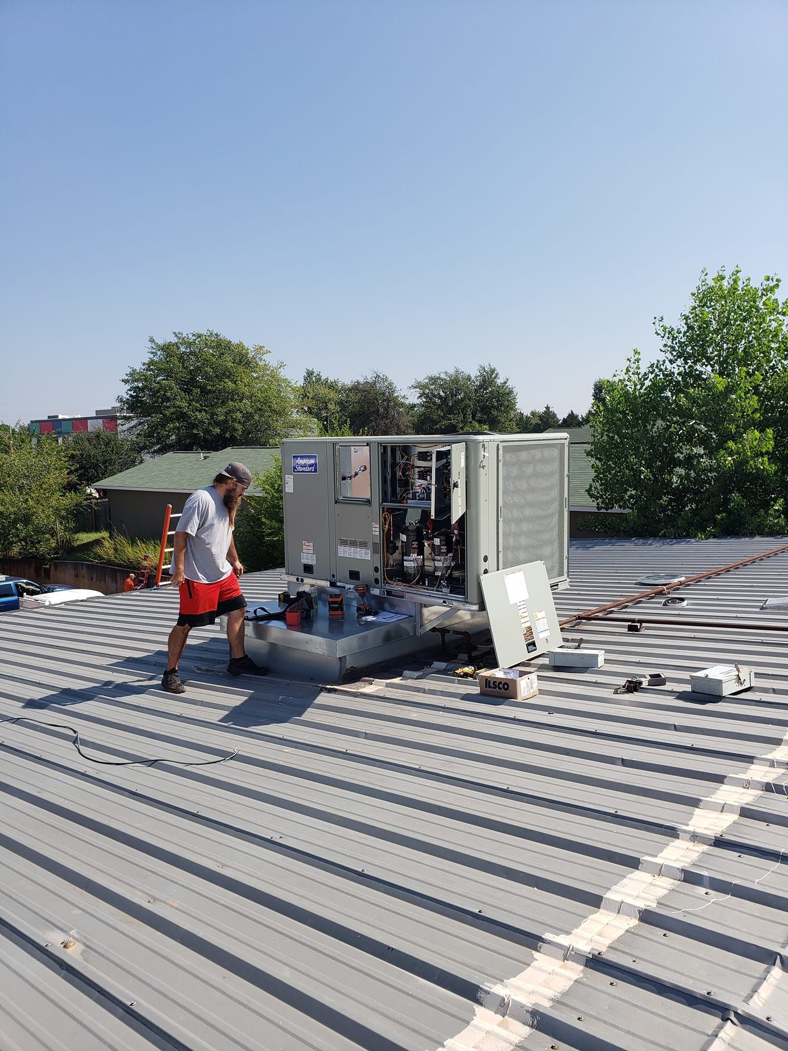 A person standing near an open HVAC unit on a metal rooftop under a blue sky.