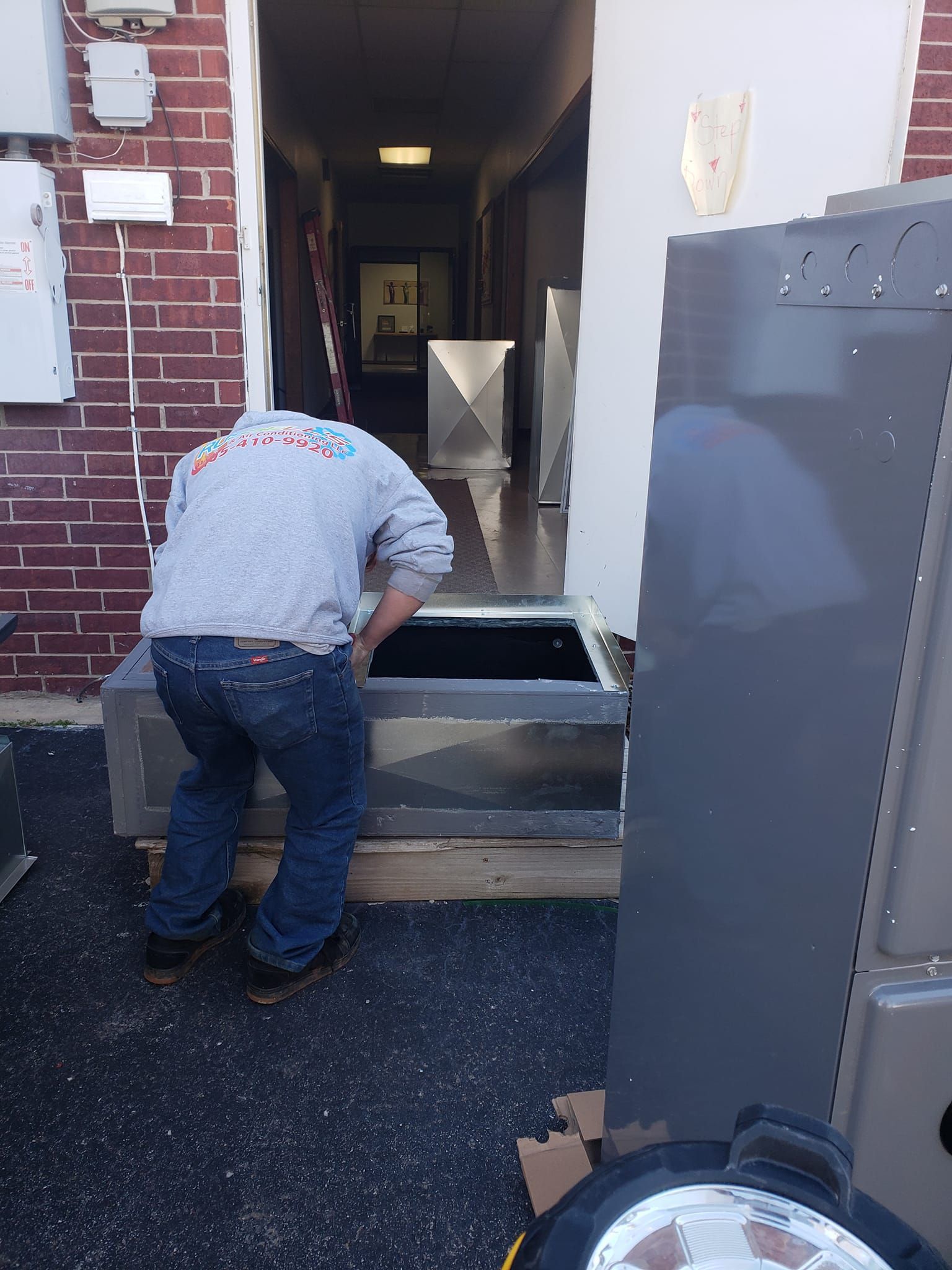 Man working on HVAC unit in brick building doorway. Metal ductwork and parts are present.