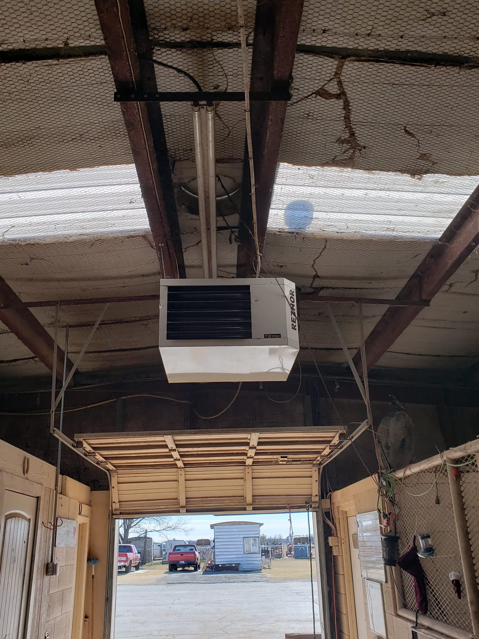 Ceiling-mounted heater inside a shed with an open doorway leading to an outdoor parking area.