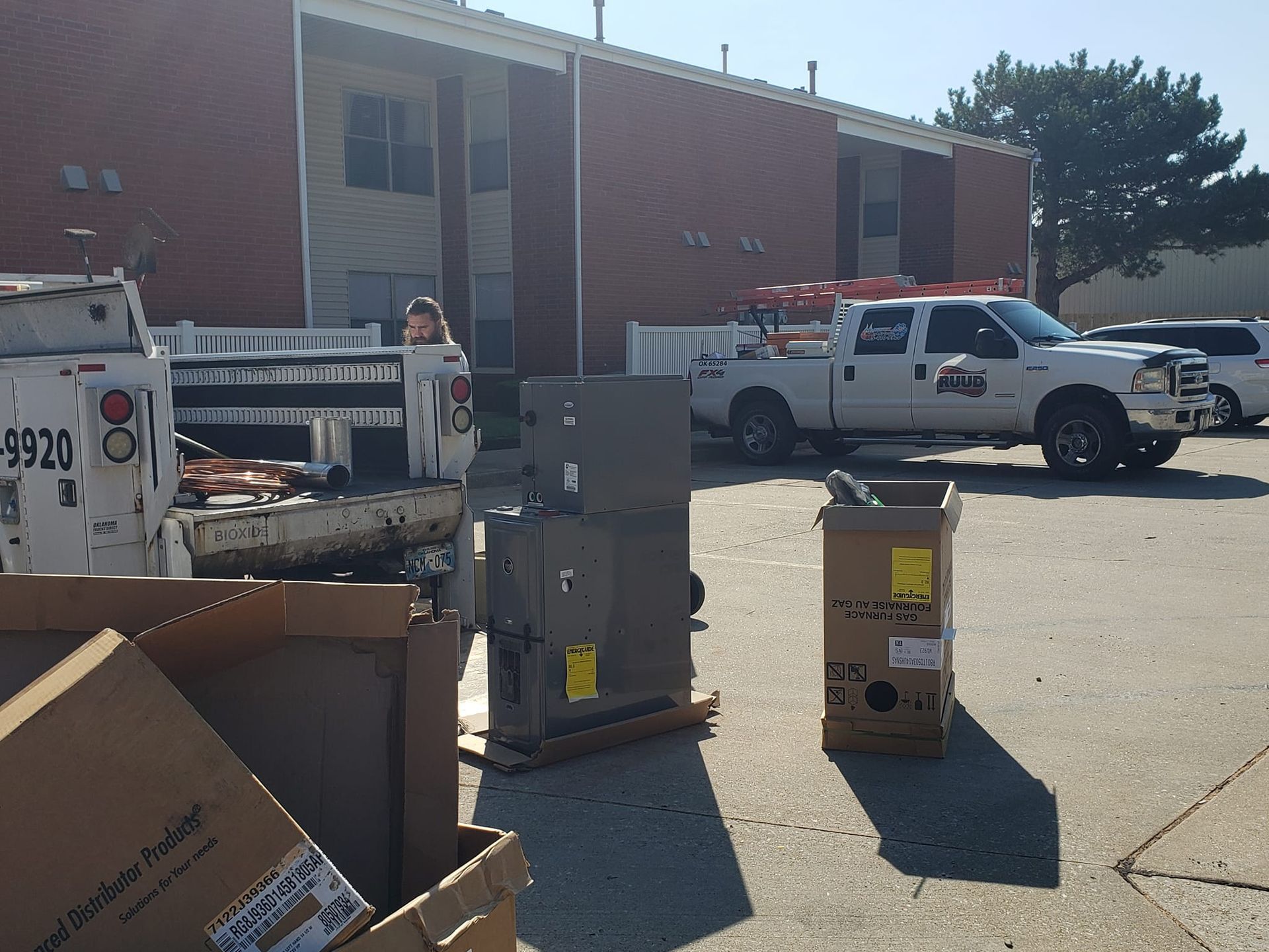 HVAC equipment and boxes outside a building with two work trucks. A person stands near a truck bed.