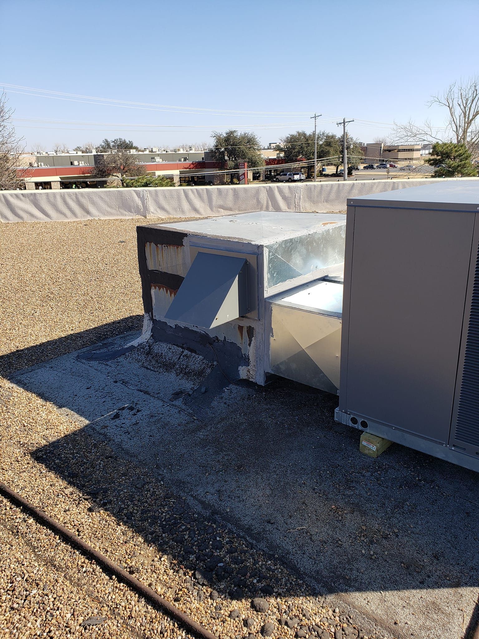 HVAC units on a flat rooftop. Silver metal with gray gravel and buildings in the background. Bright day.