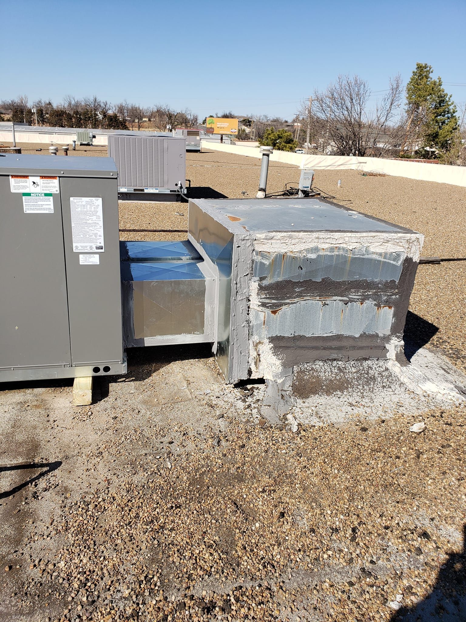 HVAC rooftop equipment; large gray unit and rusted metal box on gravel roof with sunny sky.