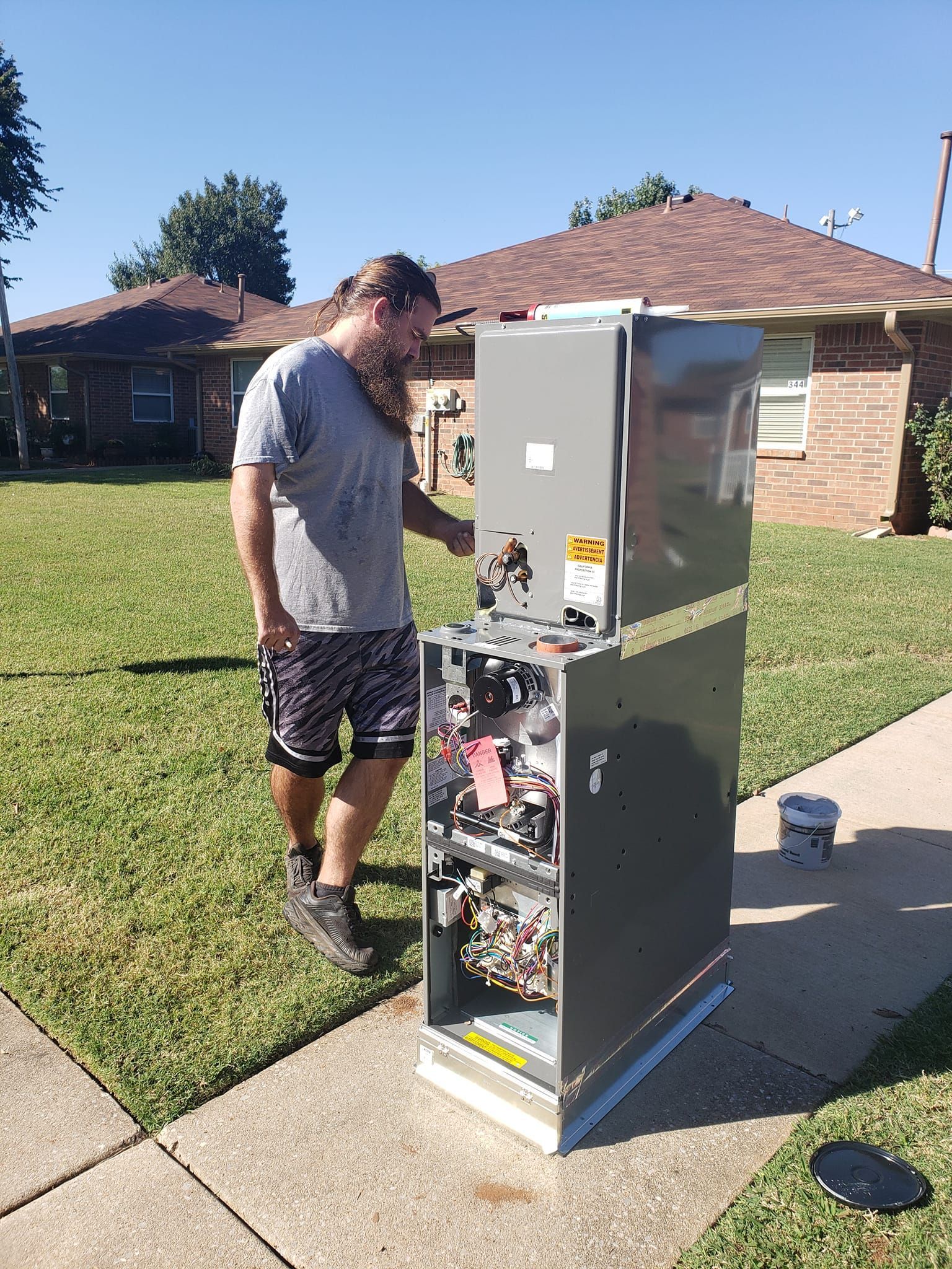 Man examines open HVAC unit on a sidewalk outside a brick home on a sunny day.