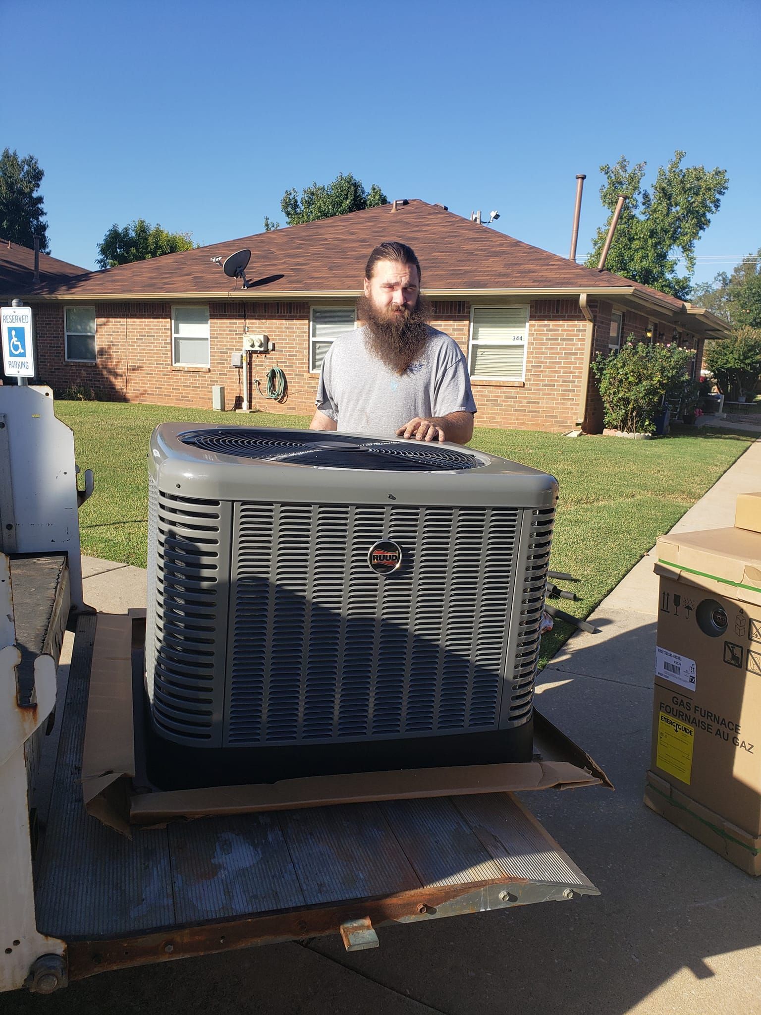 Man with a beard standing behind a large air conditioning unit on a residential driveway, sunny day.