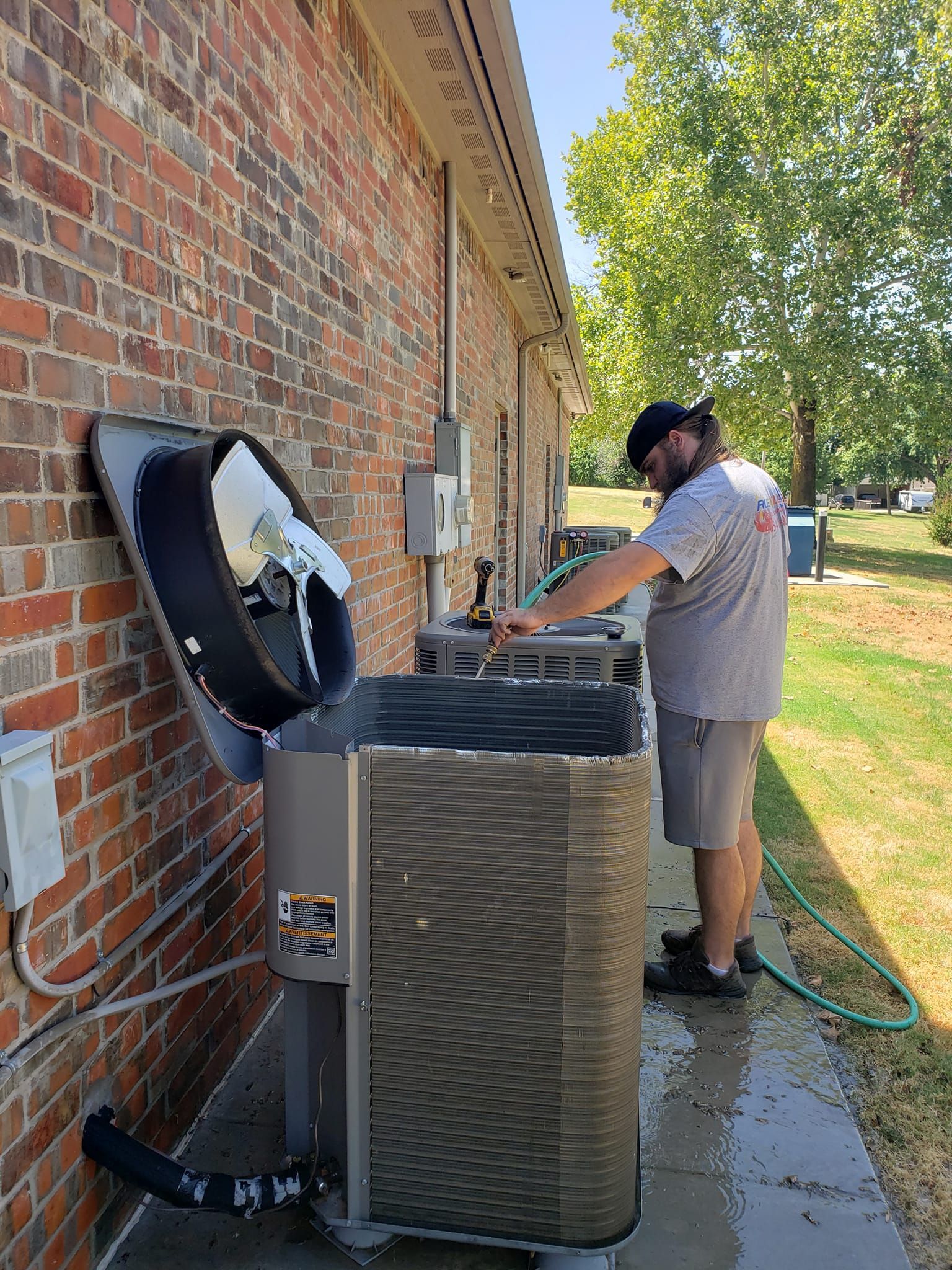 Man cleaning an air conditioner unit with a hose near a brick building.