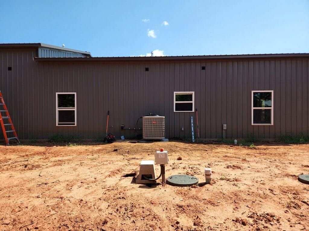 Brown building with three windows, HVAC unit, and dirt yard with septic system components.