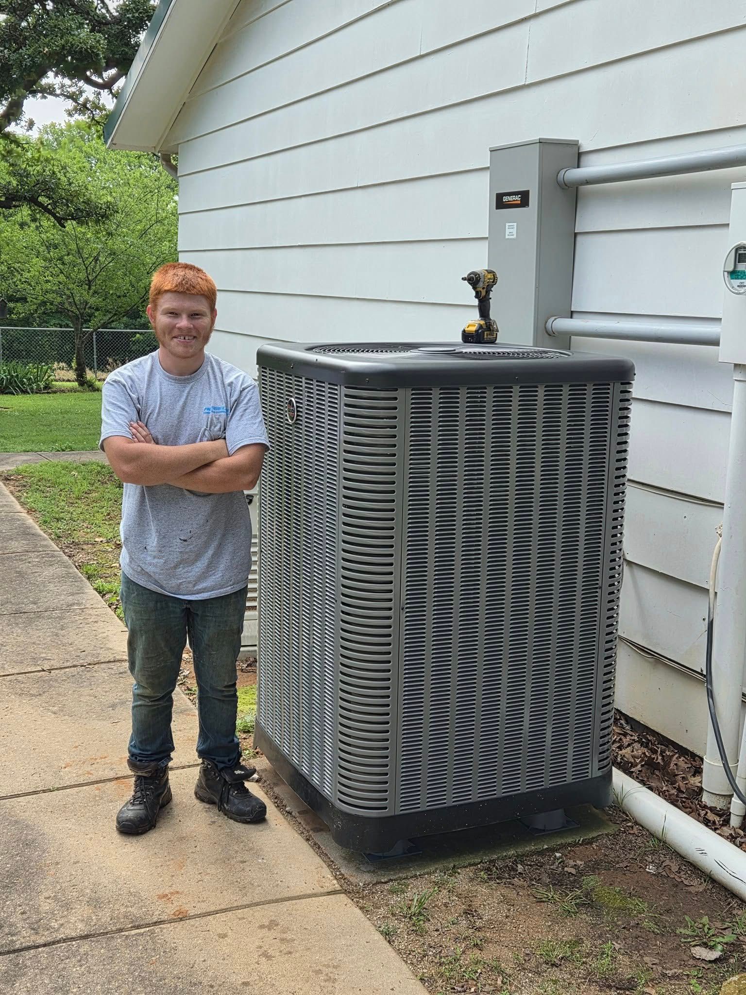 Person stands next to an air conditioning unit; arms crossed. Outdoors, near white house wall, paving stone.