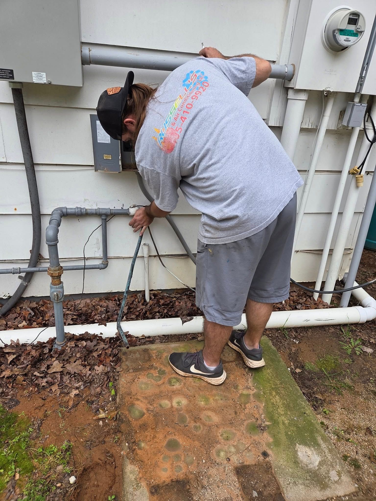 Man in gray shirt and shorts working on electrical wiring outside a building.