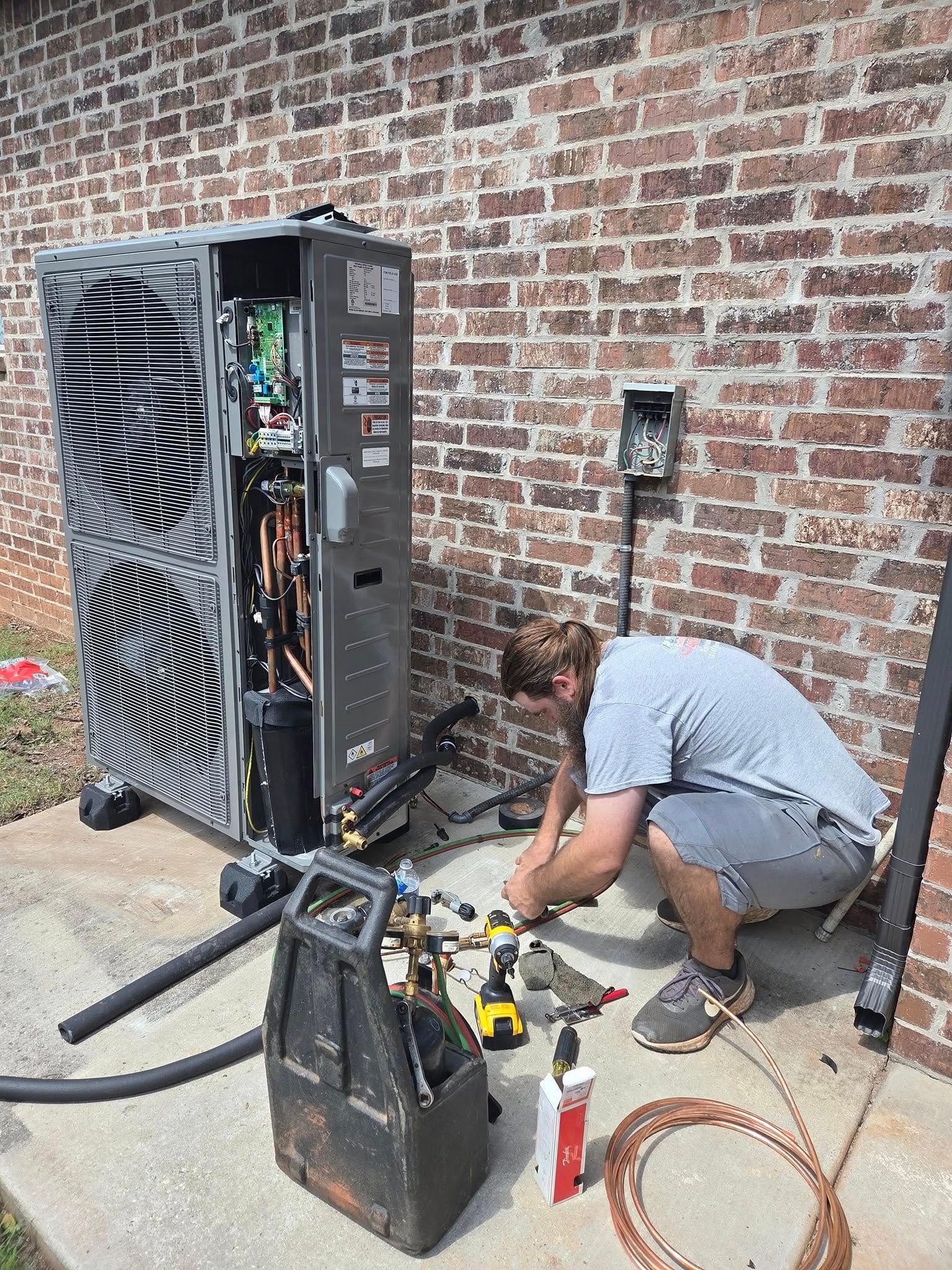 A person working on HVAC system components near a brick wall. Tools and copper tubing are in view.