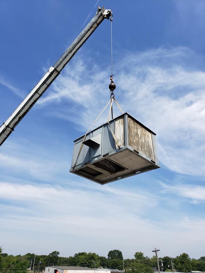 Crane lifting a large, weathered HVAC unit against a blue sky with wispy clouds.