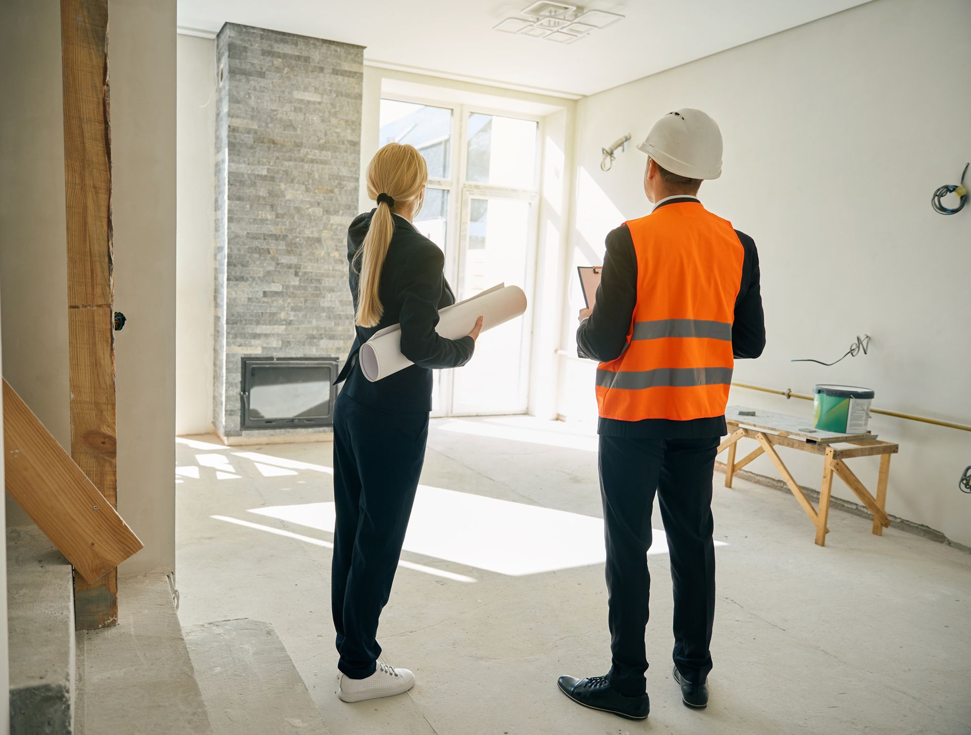 Woman and man in a construction site looking at blueprints, discussing plans near a fireplace and window.