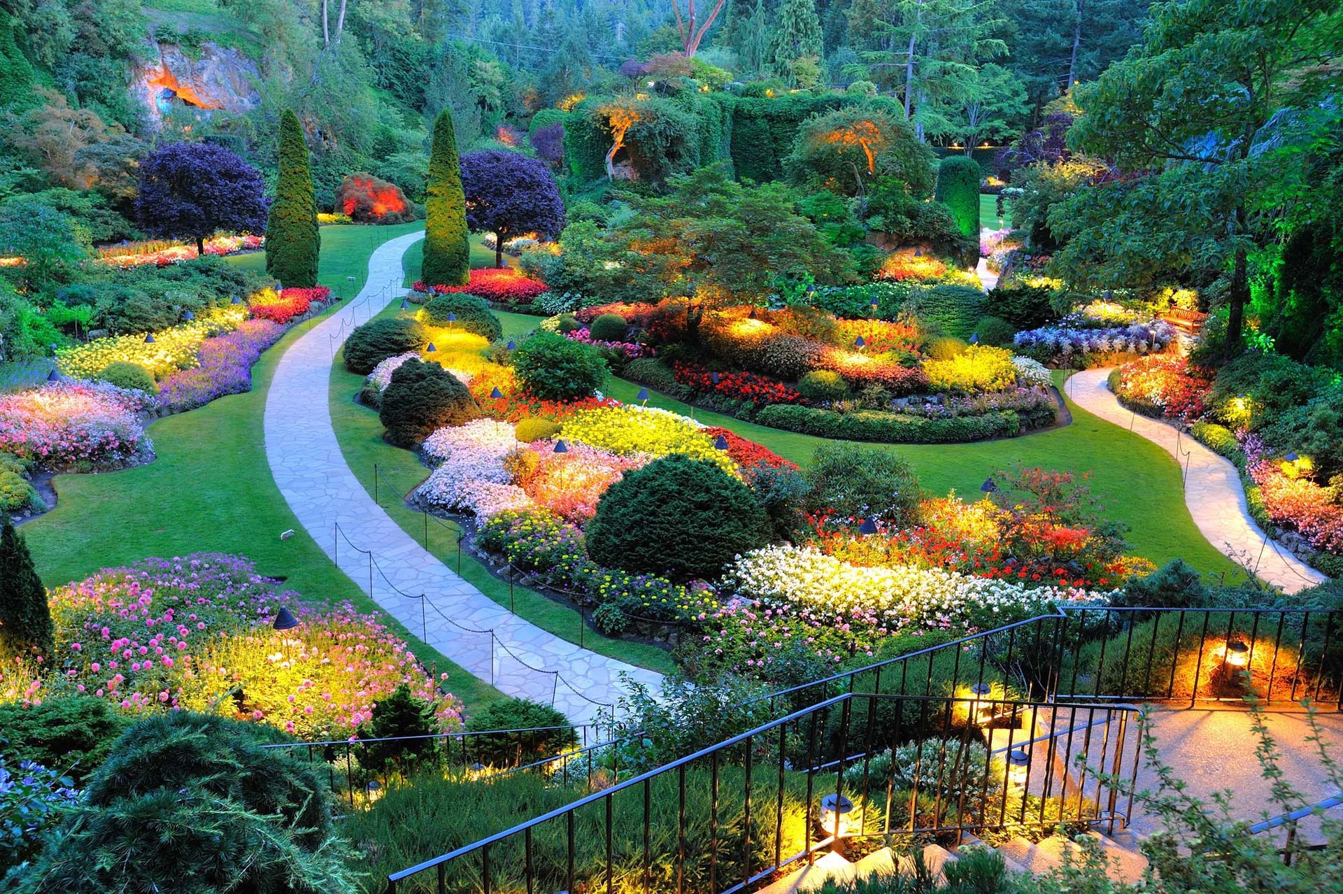 A vibrant garden path winds through colorful flowerbeds, illuminated by warm lights at dusk.