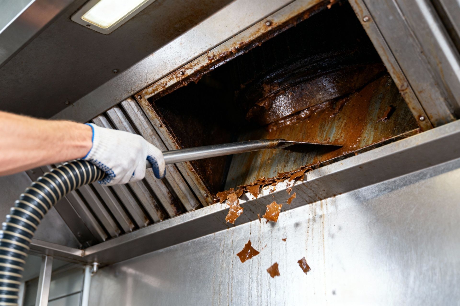 A worker wearing a glove uses a vacuum tool to clean thick, oily residue from inside a metal commercial kitchen exhaust hood.