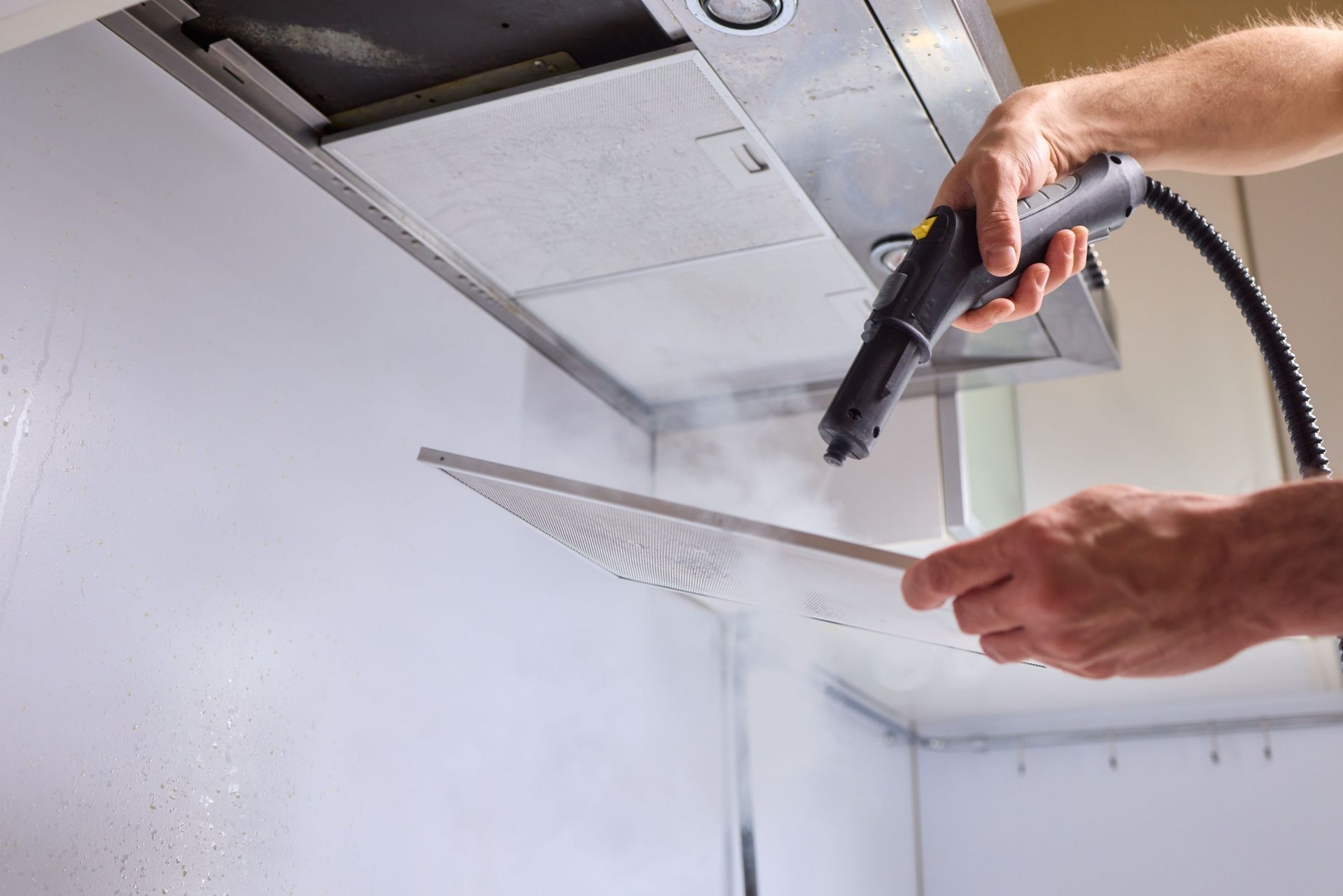 A person uses a handheld steam cleaner to sanitize a metal kitchen range hood filter.