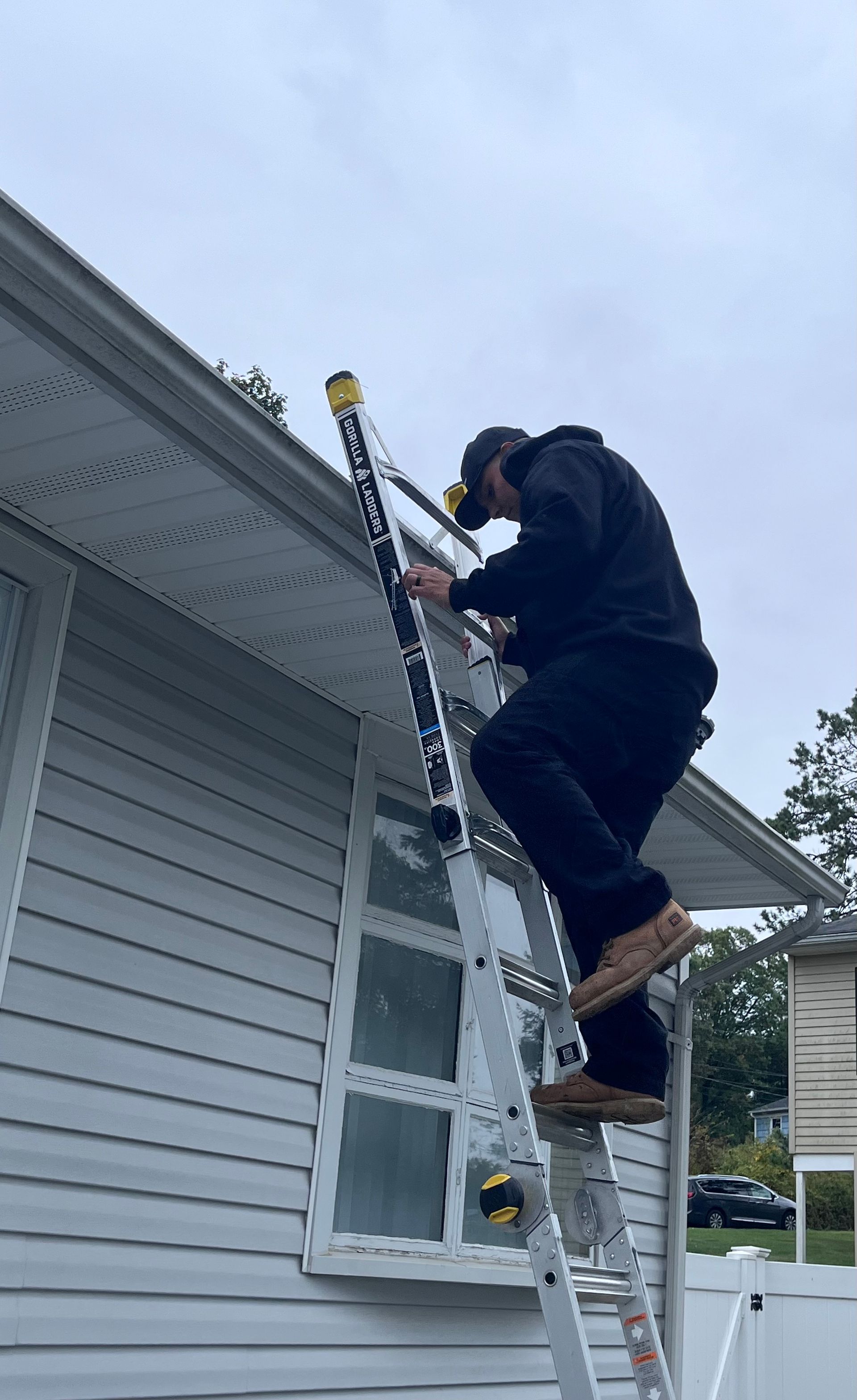 Man on a ladder, cleaning gutters on a house with light gray siding under an overcast sky.