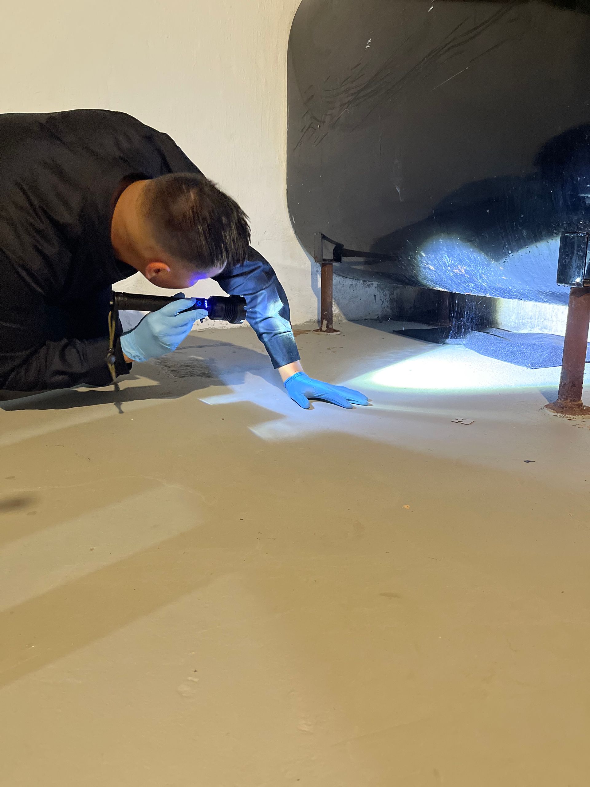 Person kneeling, wearing gloves, shining a flashlight on a floor with debris. Dark, enclosed space.