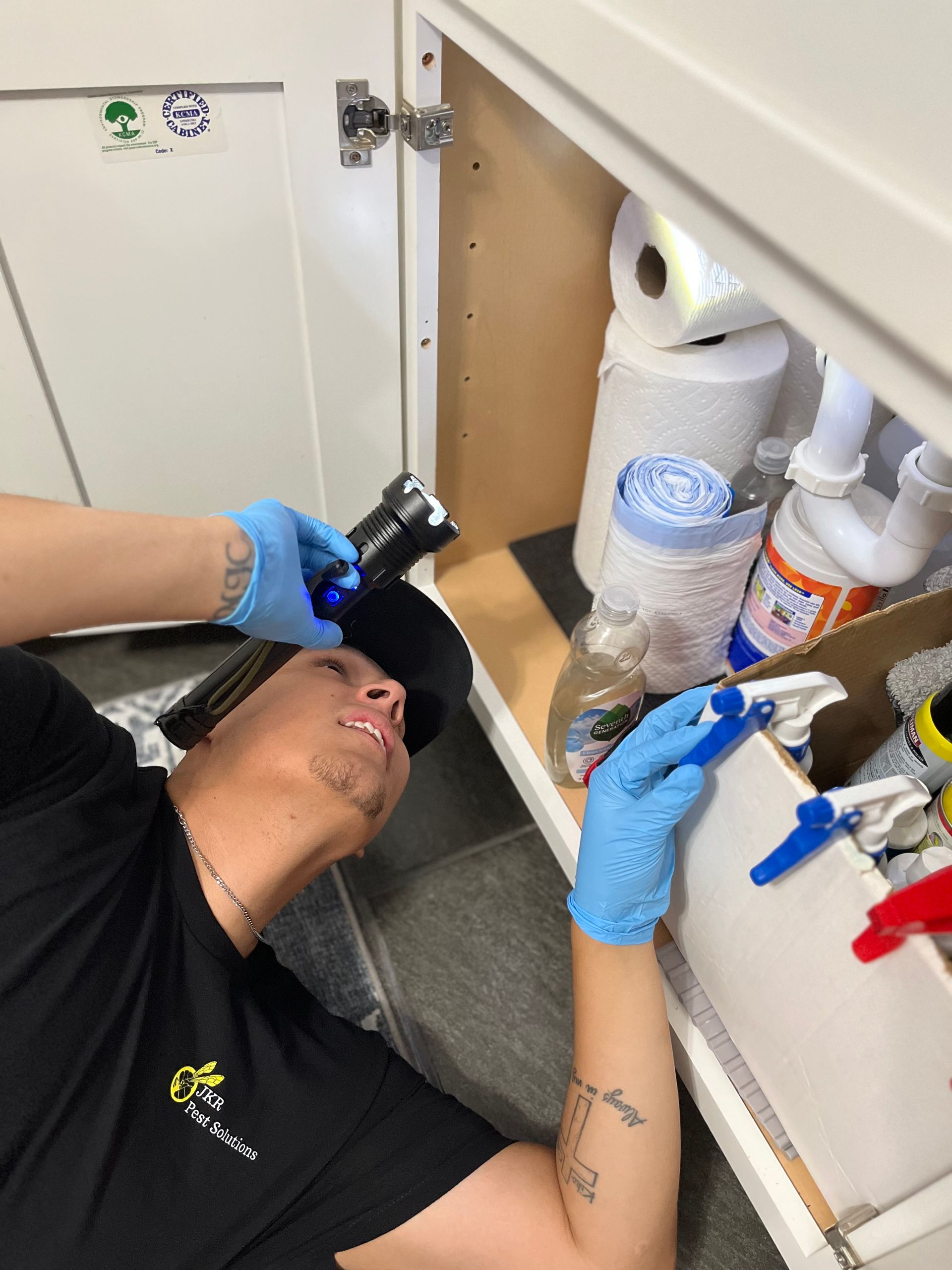Plumber in blue gloves examines pipes under a sink, using a flashlight.