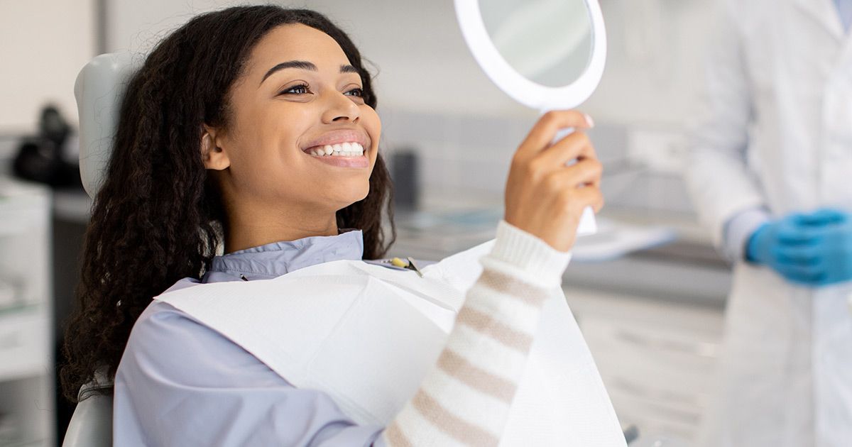 A woman is sitting in a dental chair looking at her teeth in a mirror.