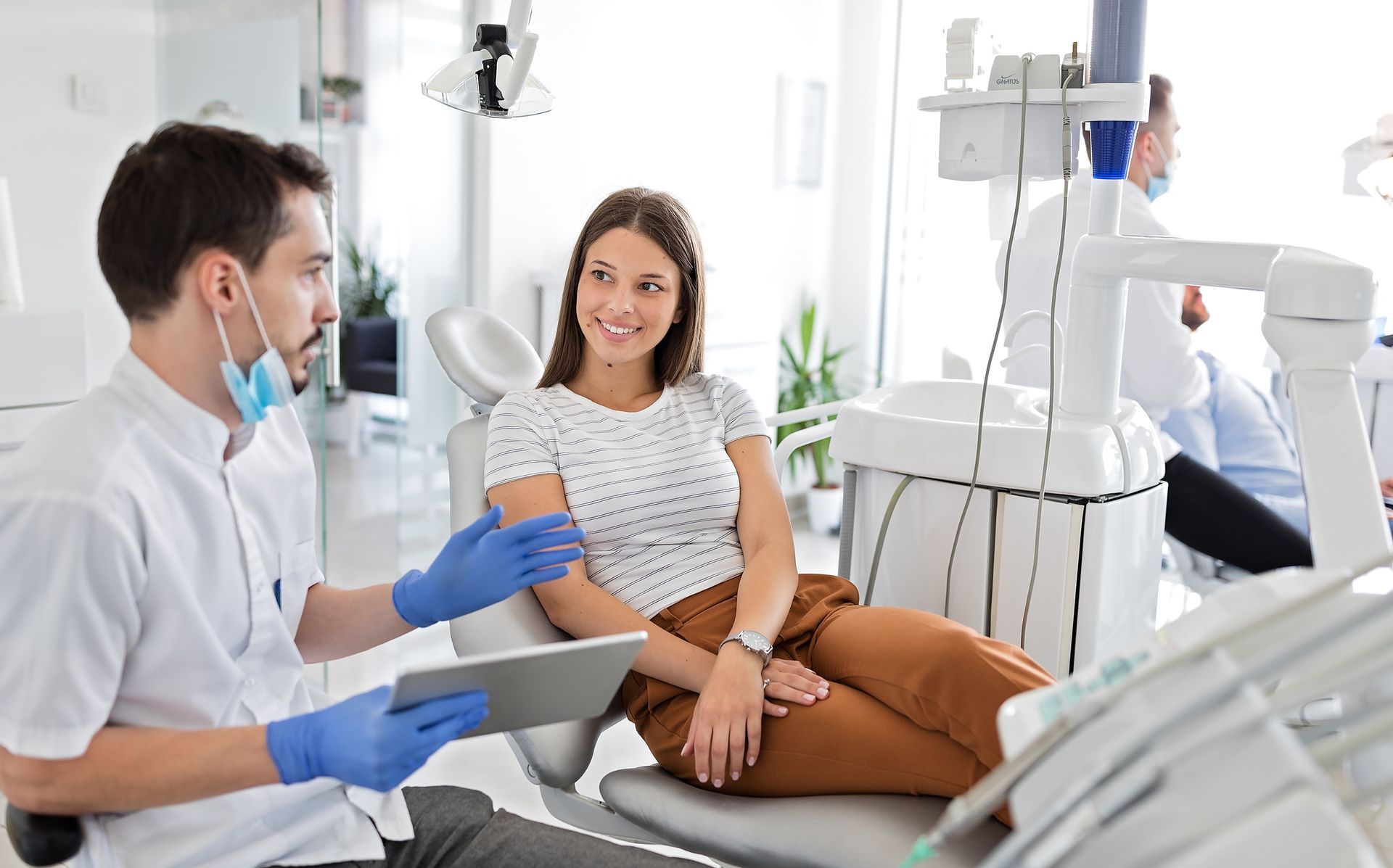 Dentist in mask and gloves consults with patient in dental chair; bright office setting.