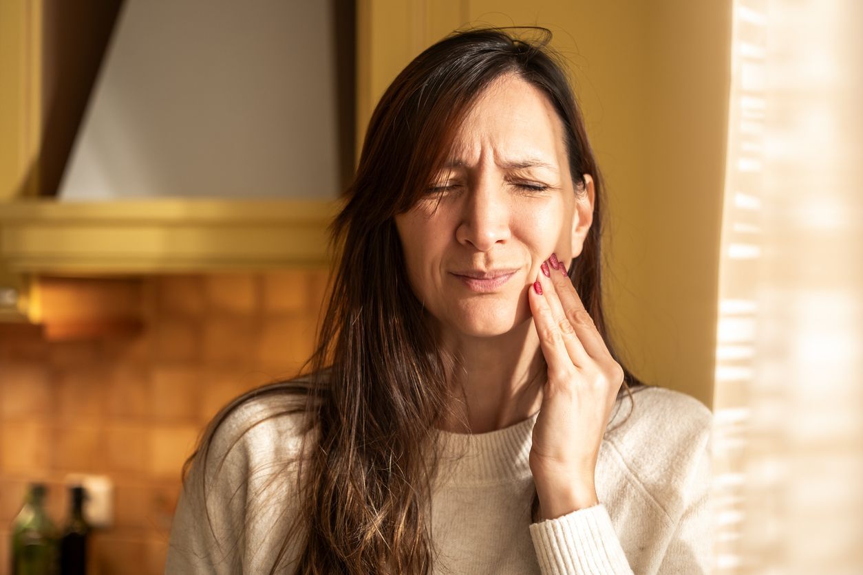 Woman with her hand on her cheek, grimacing in pain. She is indoors with a blurred kitchen background.