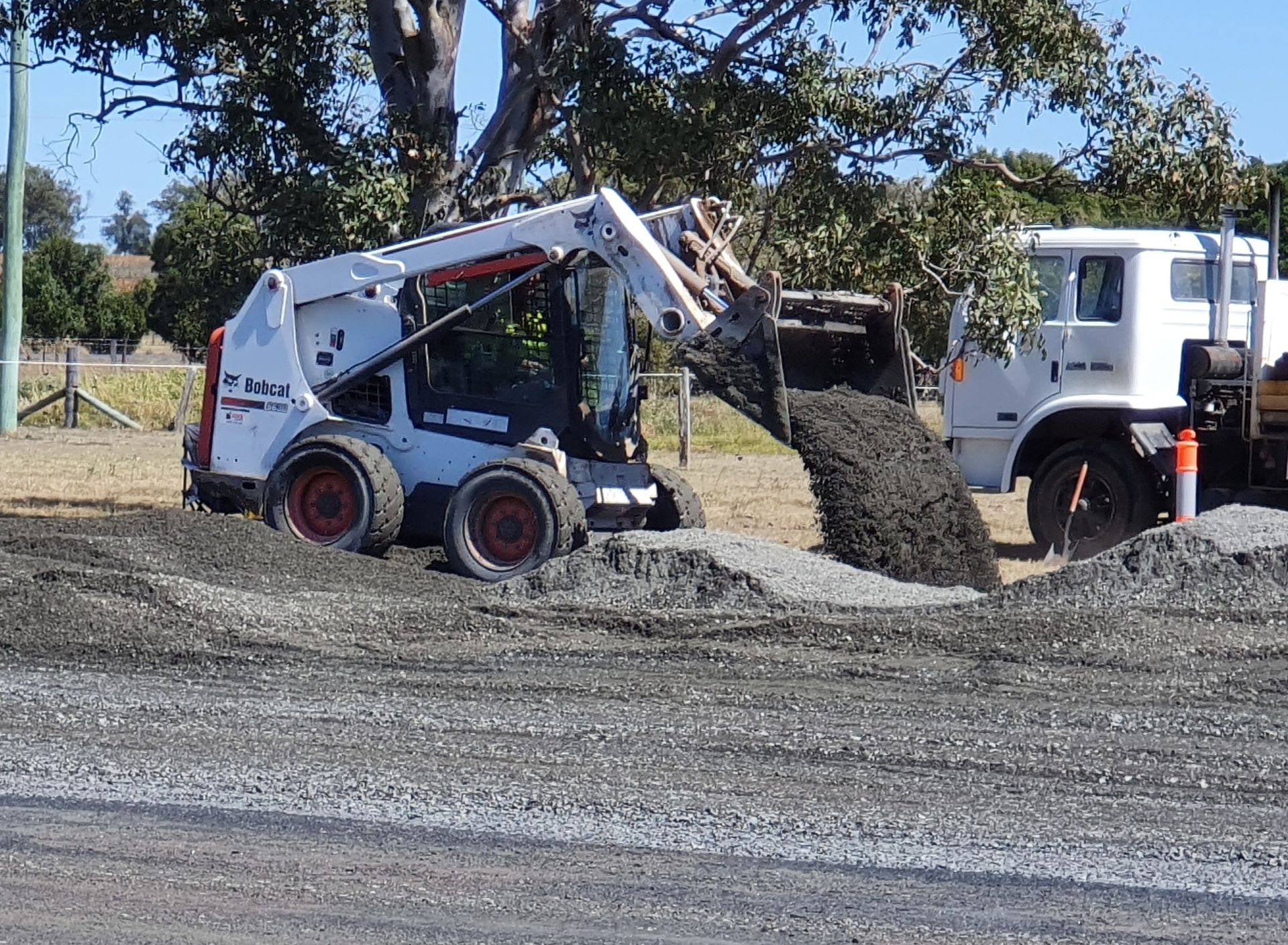 A Bobcat Skid Steer Dumping Gravel on a Road, Beside a White Truck — Snell Earthworx In Maryborough, QLD