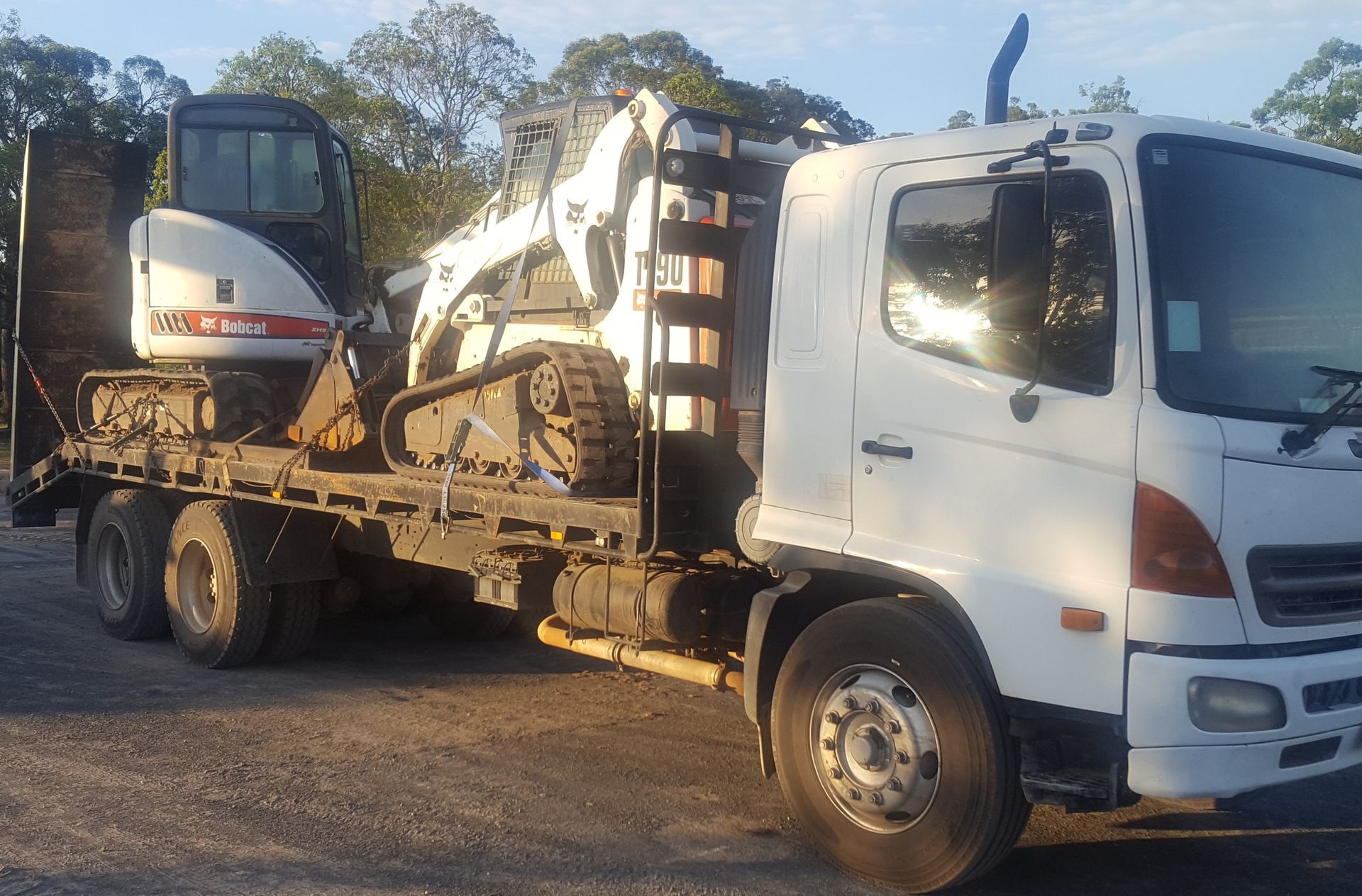 White truck with a Bobcat excavator loaded on the flatbed, outdoors. — Snell Earthworx In Craignish, QLD