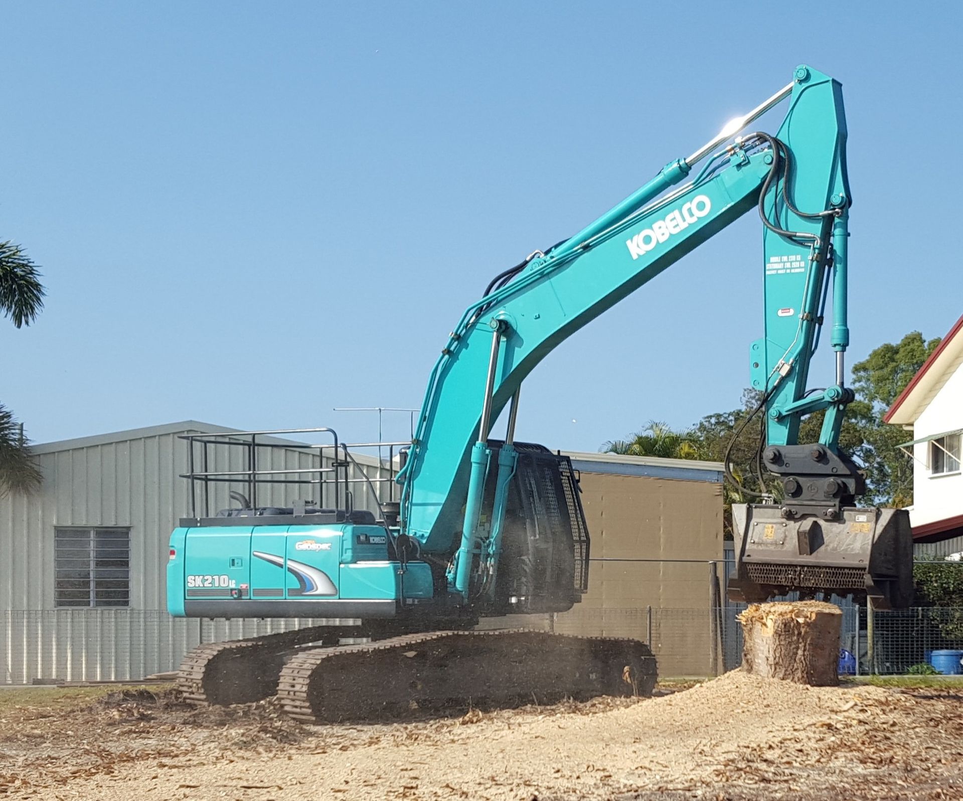 A teal excavator working on a stump outdoors on a sunny day. — Snell Earthworx In Hervey Bay, QLD