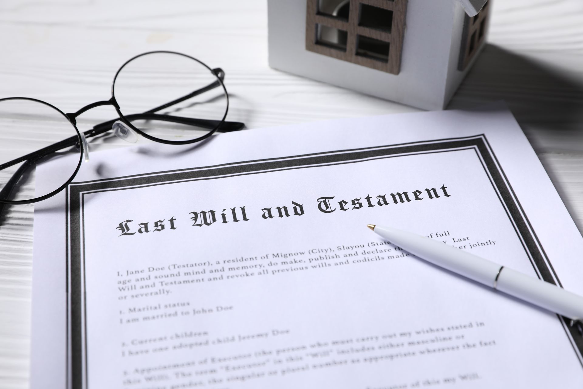 A Last Will and Testament document with a pen, eyeglasses, and a miniature house model on a white wooden surface. A Last Will and Testament document with a pen, eyeglasses, and a miniature house model on a white wooden surface.