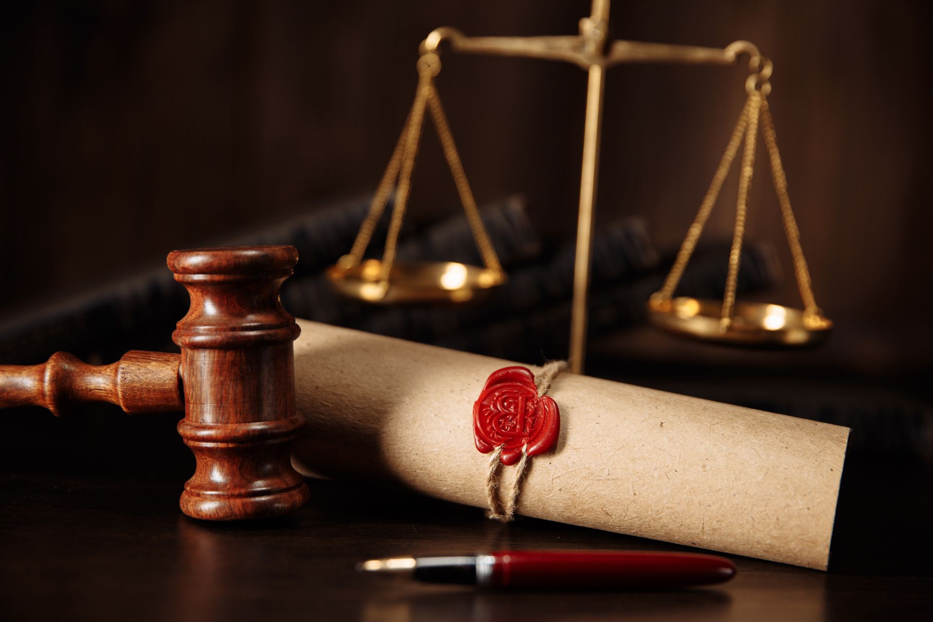 A wooden gavel, a sealed scroll, and a fountain pen resting in front of a pair of golden scales of justice.