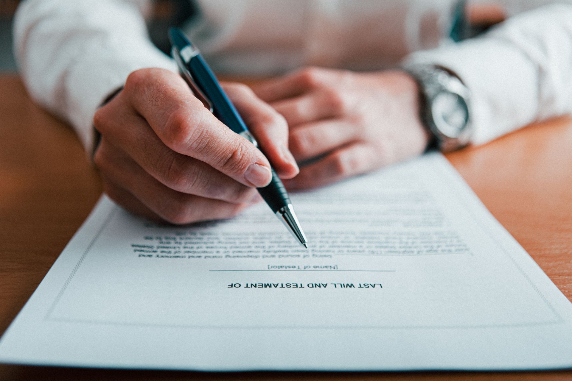 A close-up of a person in a white shirt signing a Last Will and Testament document with a pen at a wooden desk.
