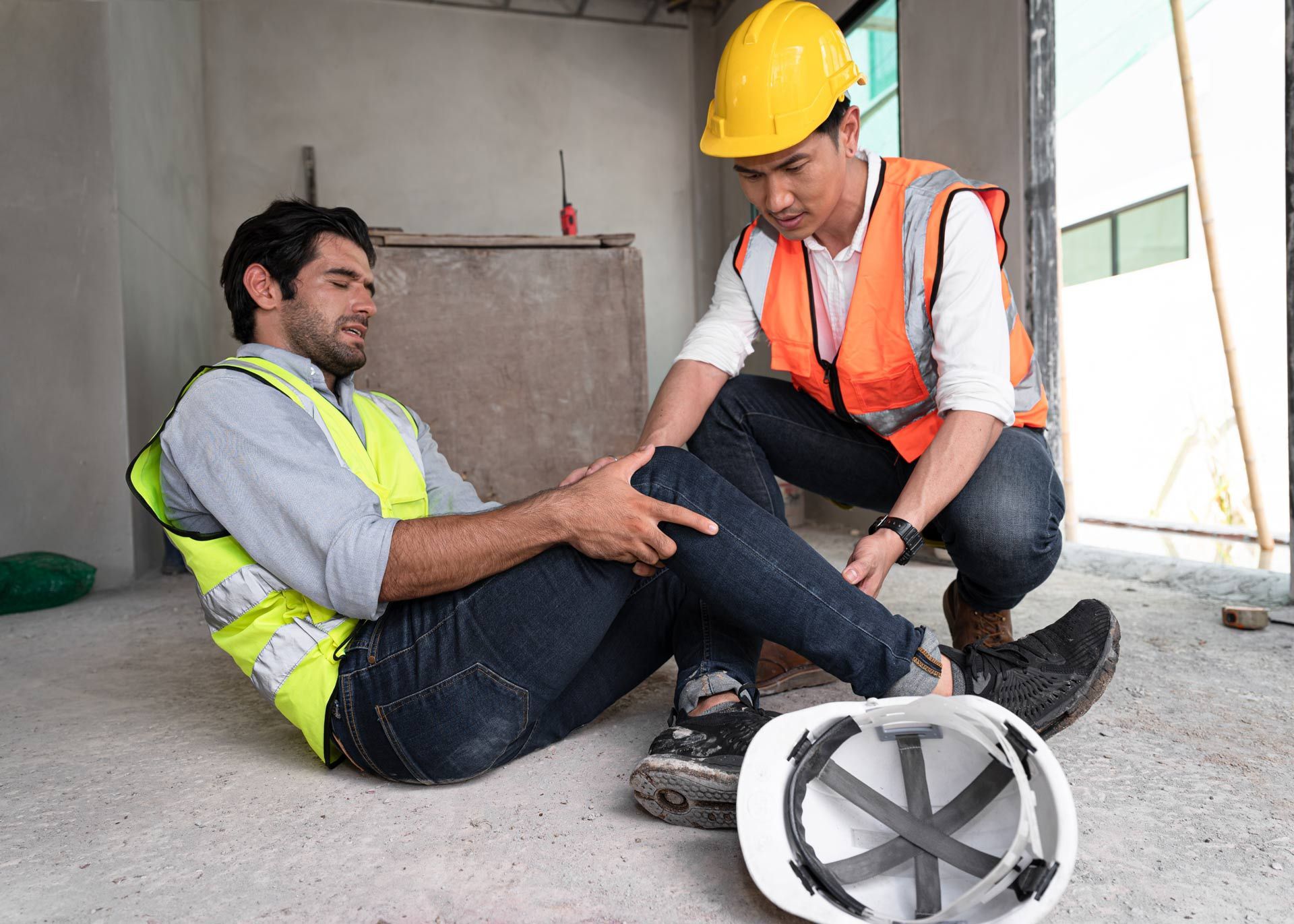 A male engineer kneels in pain while another helps him to hold his leg, inside a construction site.