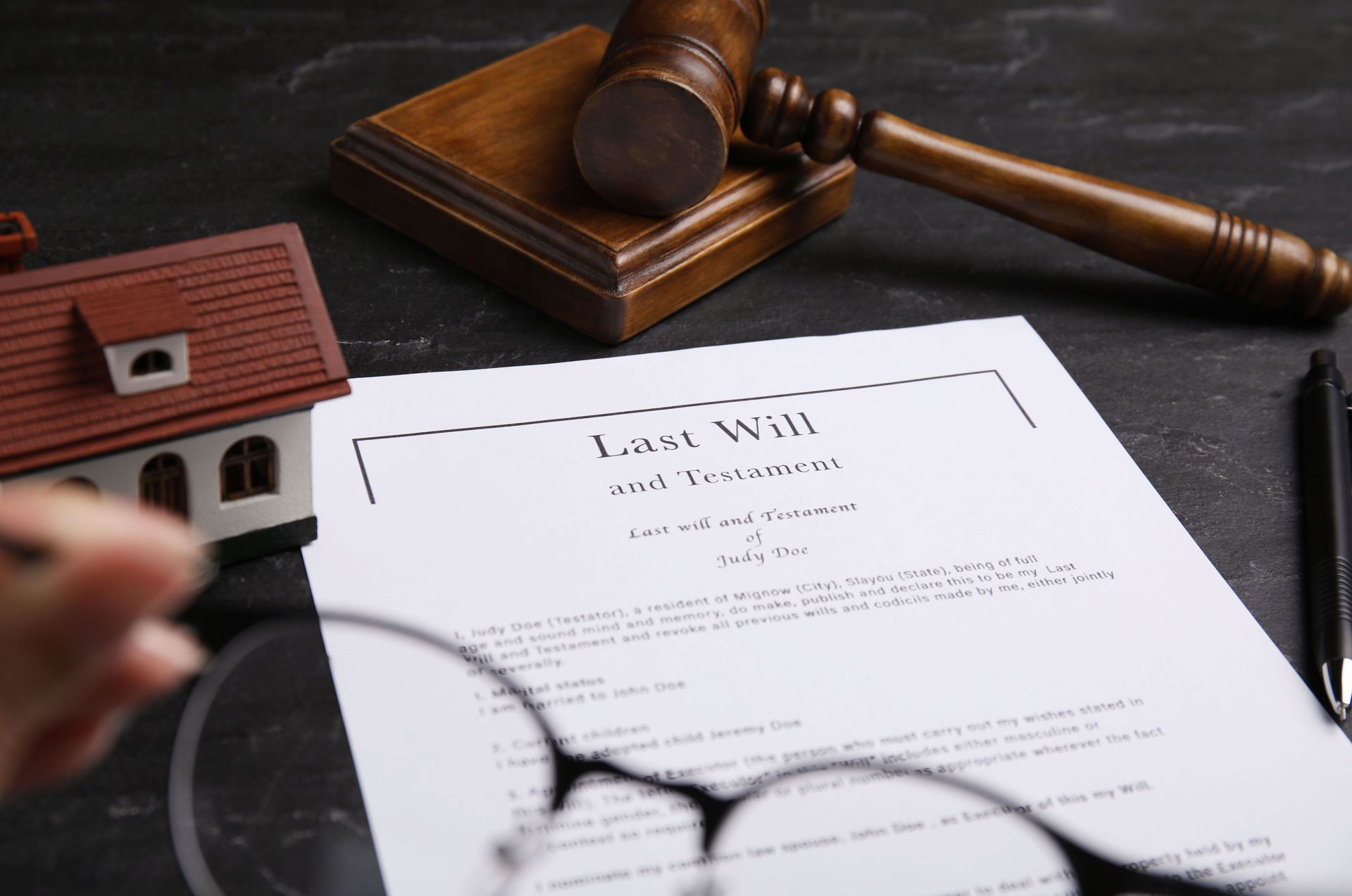 A Last Will and Testament document sits on a dark table next to a model house, a wooden gavel, and blurred eyeglasses.