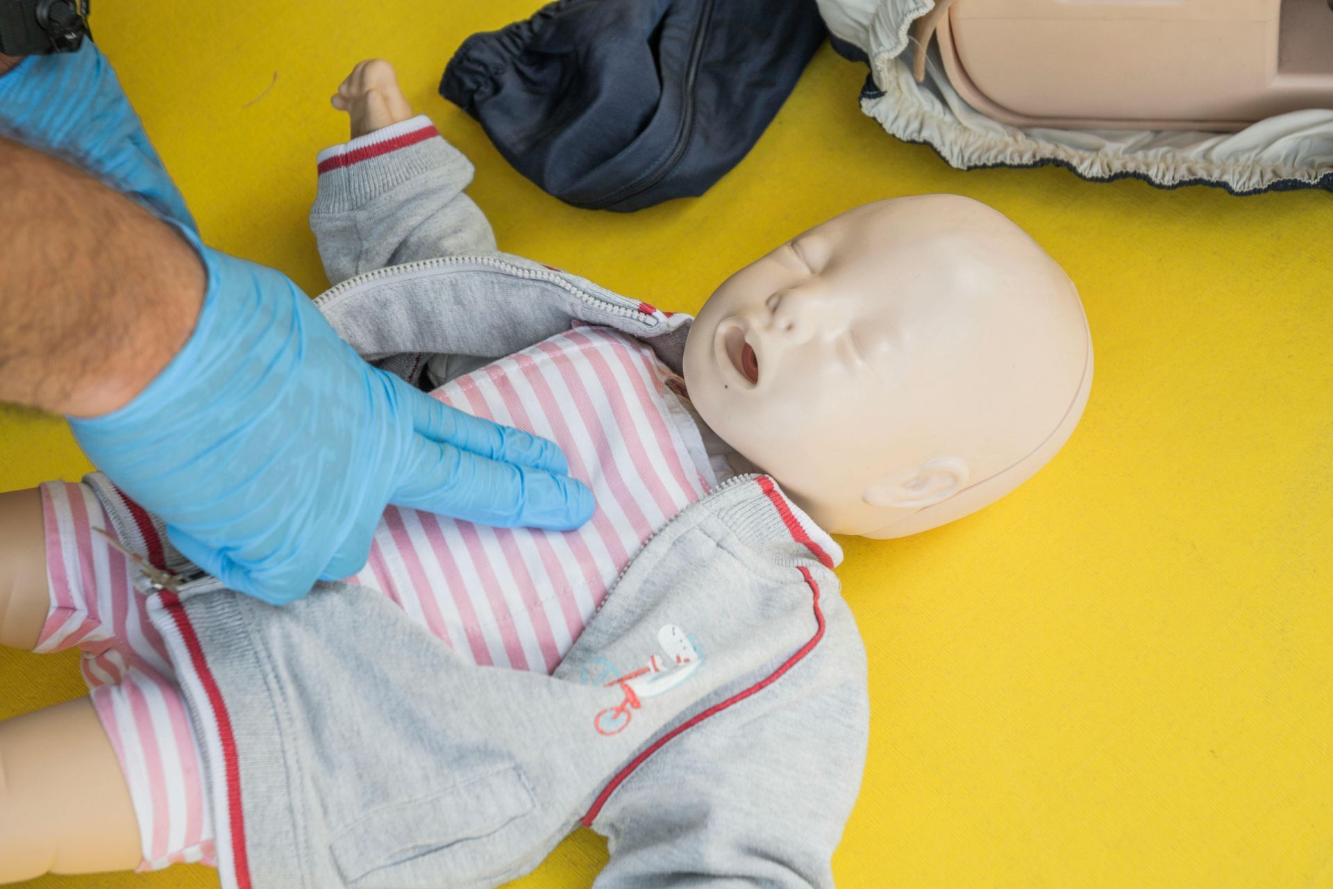 A person in blue gloves demonstrates infant CPR on a mannequin. The mannequin lies on a yellow surface, wearing a striped shirt and gray sweater.
