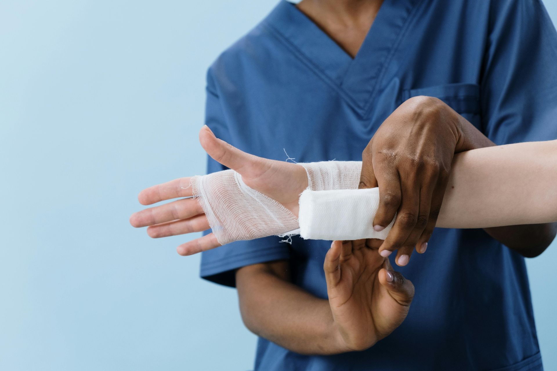 A healthcare worker in blue scrubs wraps a bandage around a patient's wrist. Dark-skinned hands attend to a light-skinned arm.