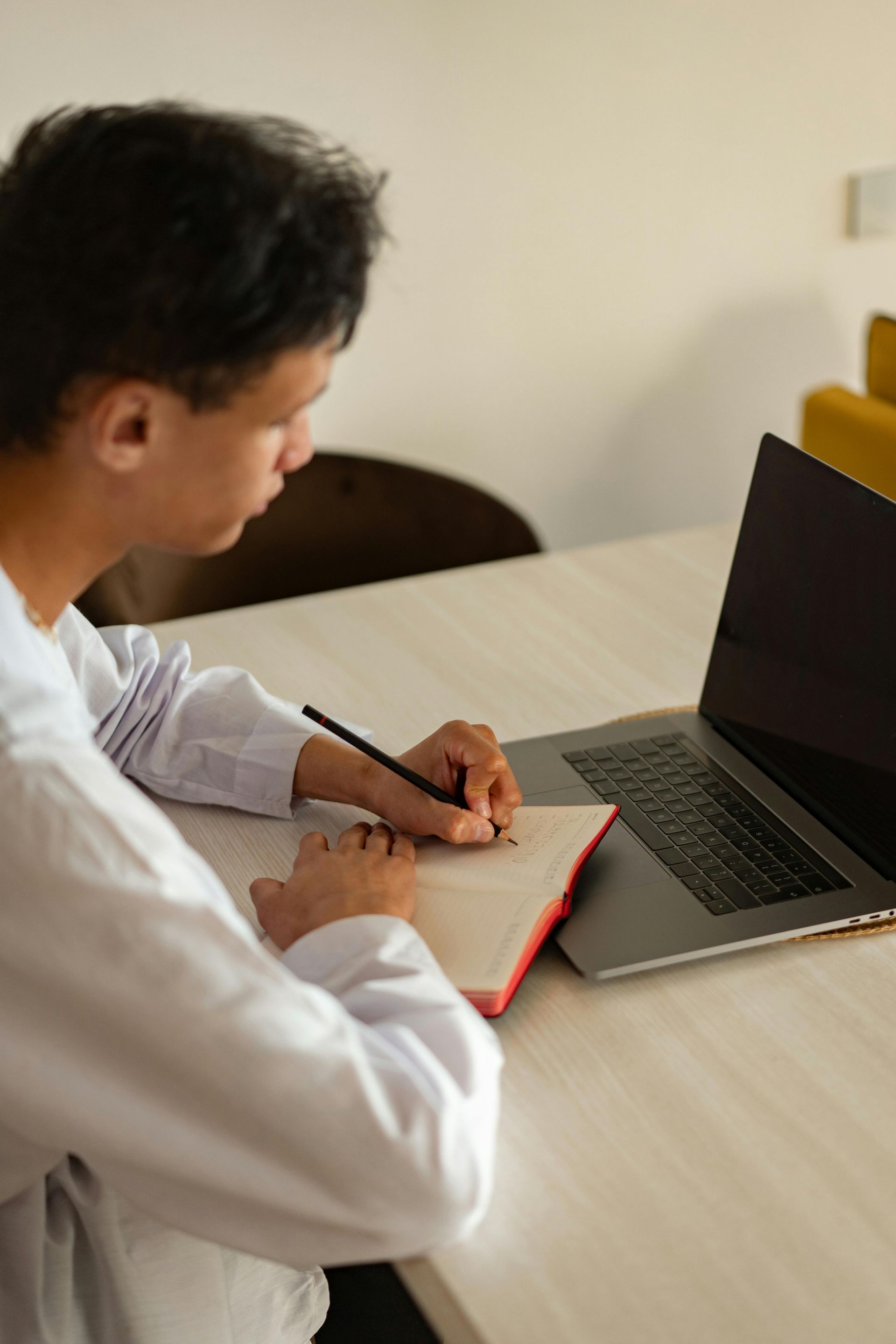 Person in white shirt writing in notebook at a desk with a laptop; interior setting.