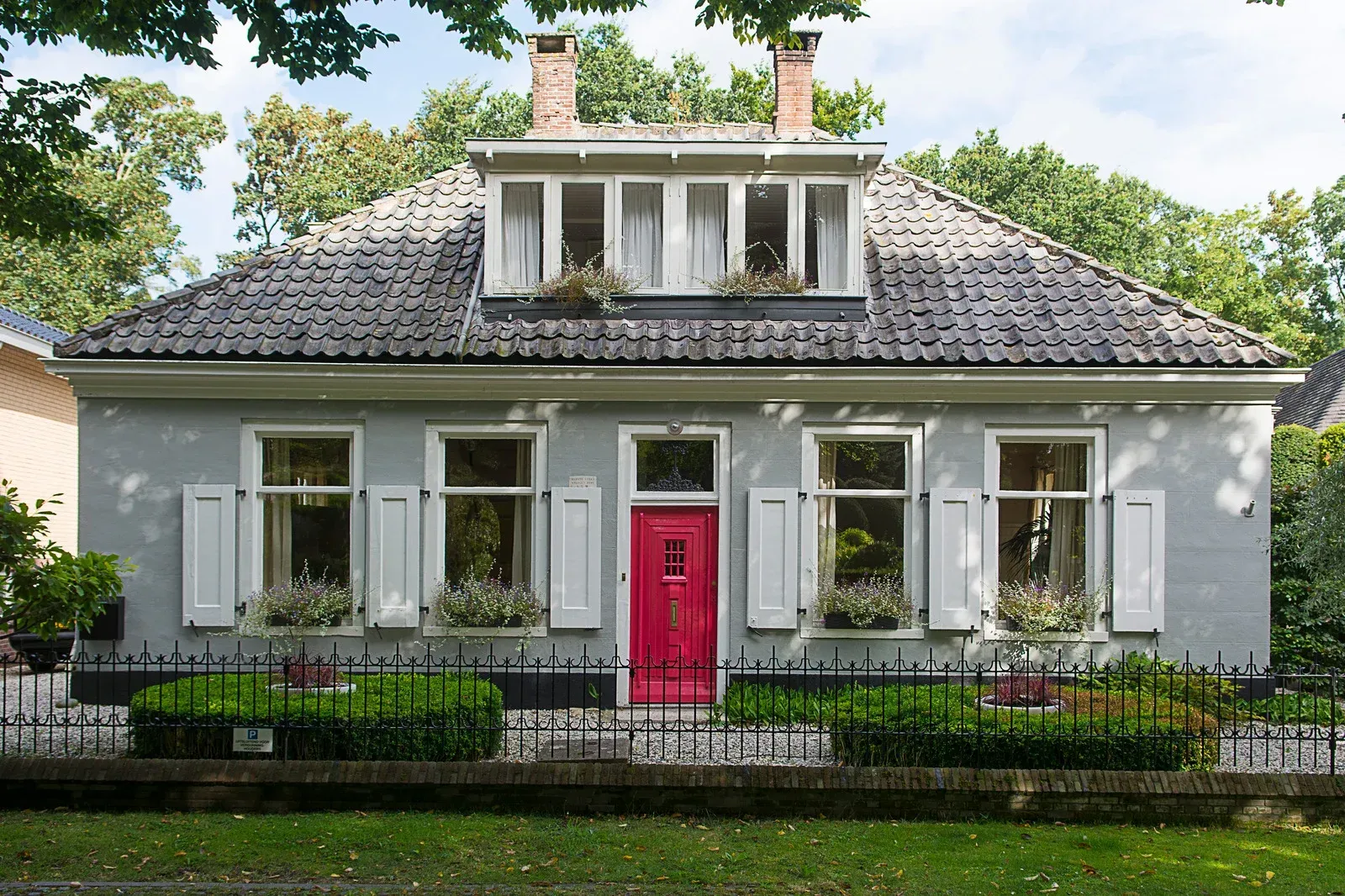 Gray house with red door, white shutters, small dormer windows, and a black fence.