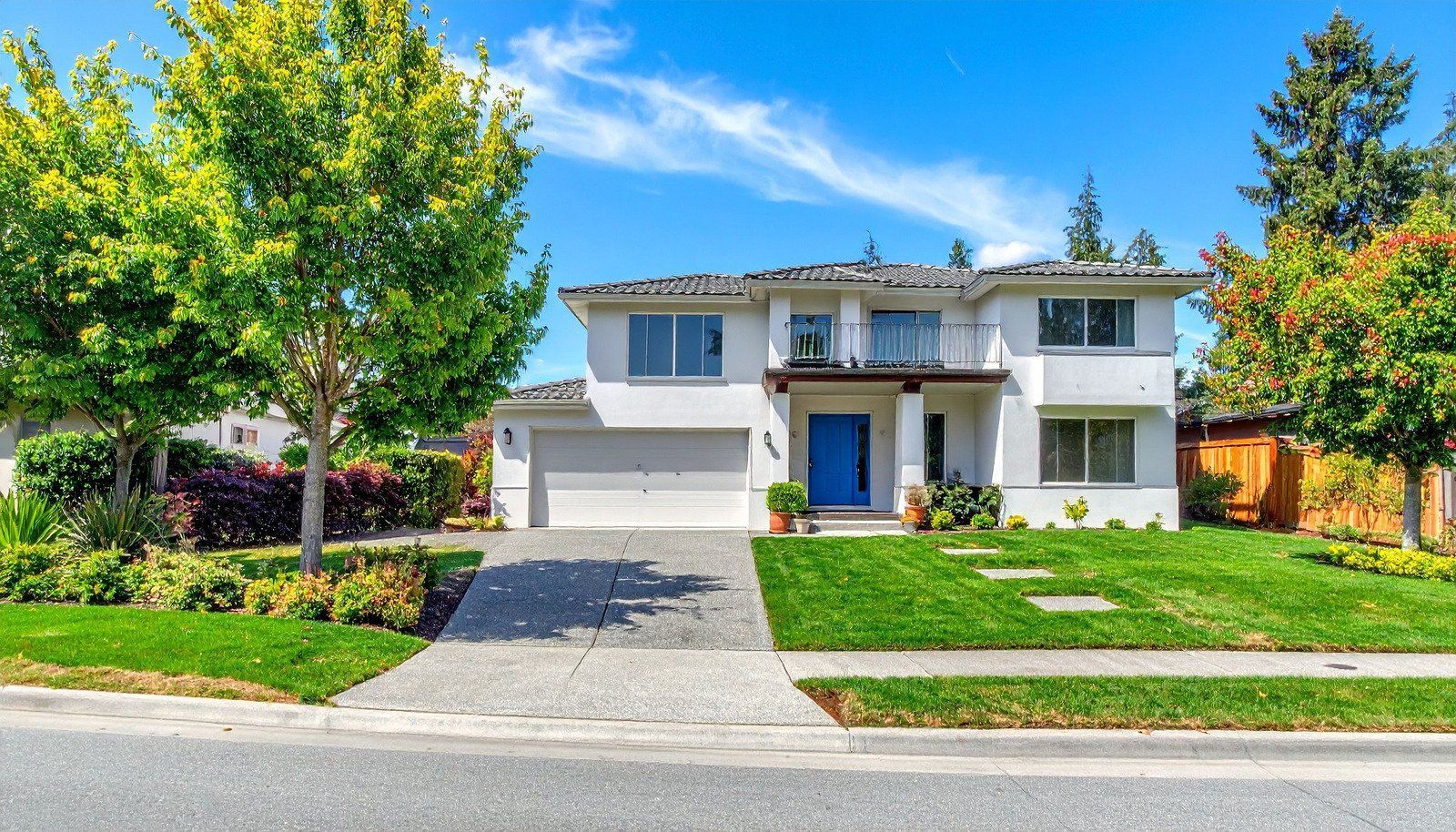 Two-story white house with blue door, green lawn, trees, and driveway under a blue sky.
