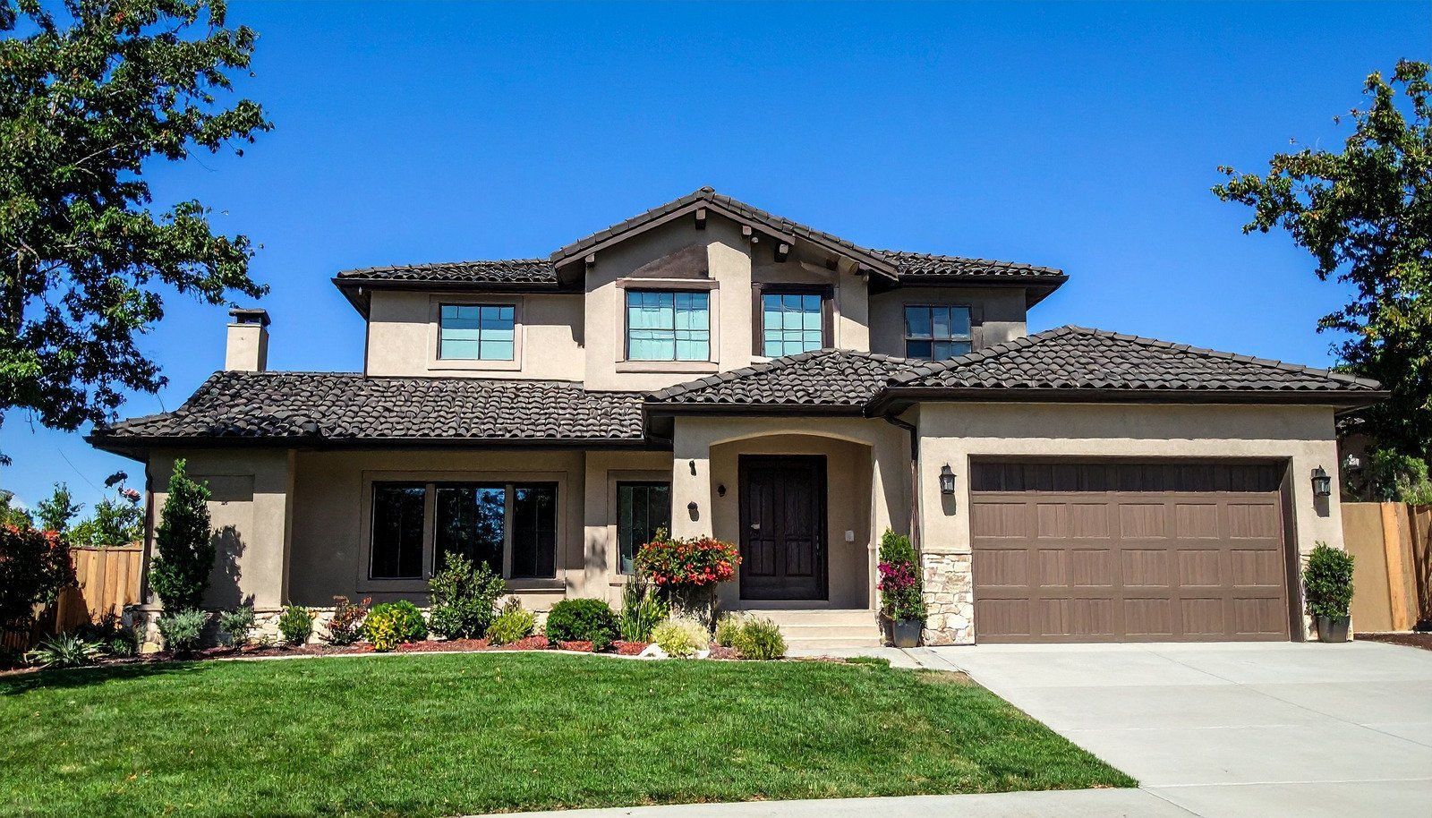 Two-story beige house with brown tile roof, garage, and green lawn under a clear blue sky.