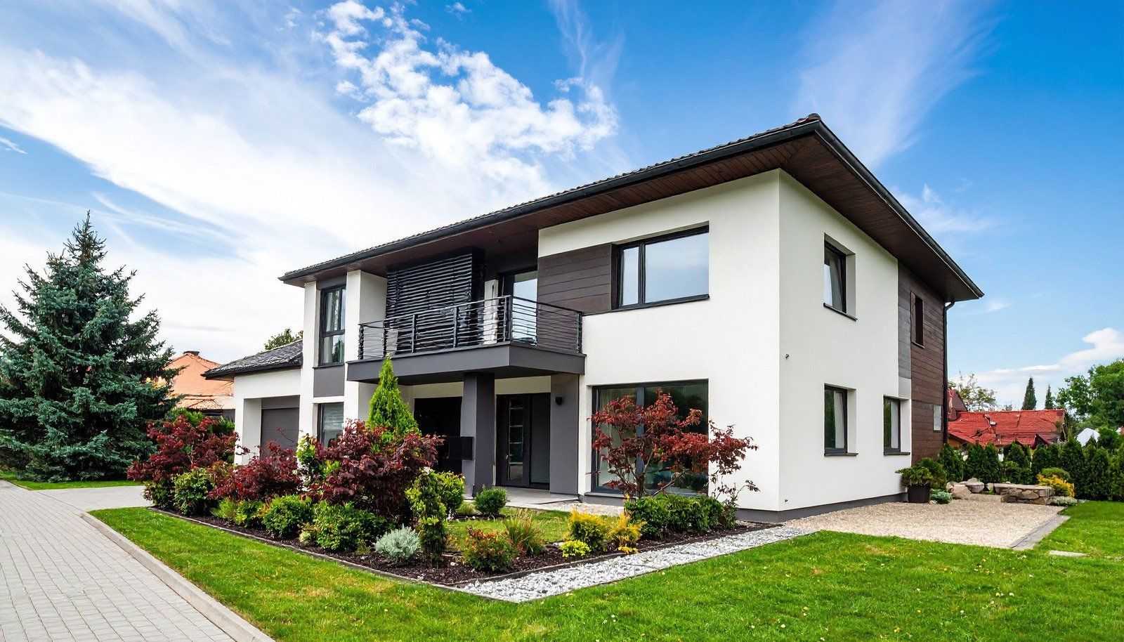 Modern two-story house with white and brown exterior, balcony, and landscaped yard under a blue sky.