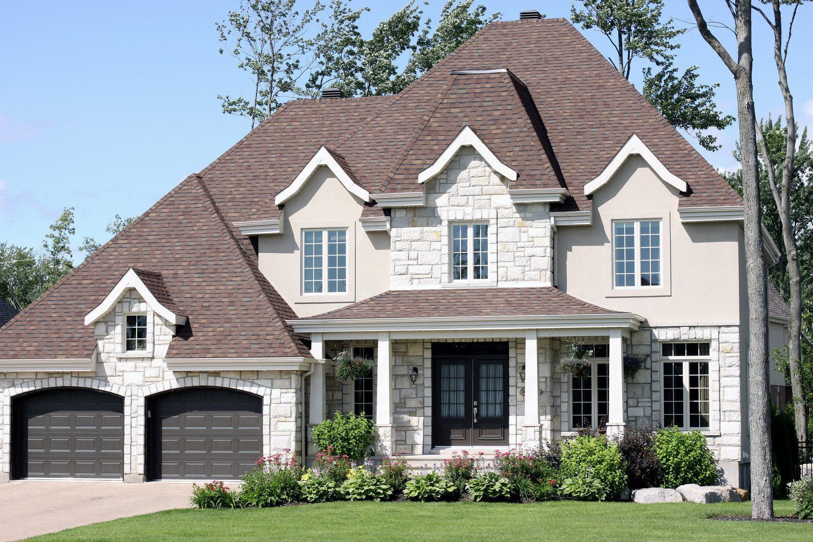 Two-story house with stone facade, dark brown roof, two-car garage, and front porch. Green lawn and shrubbery.