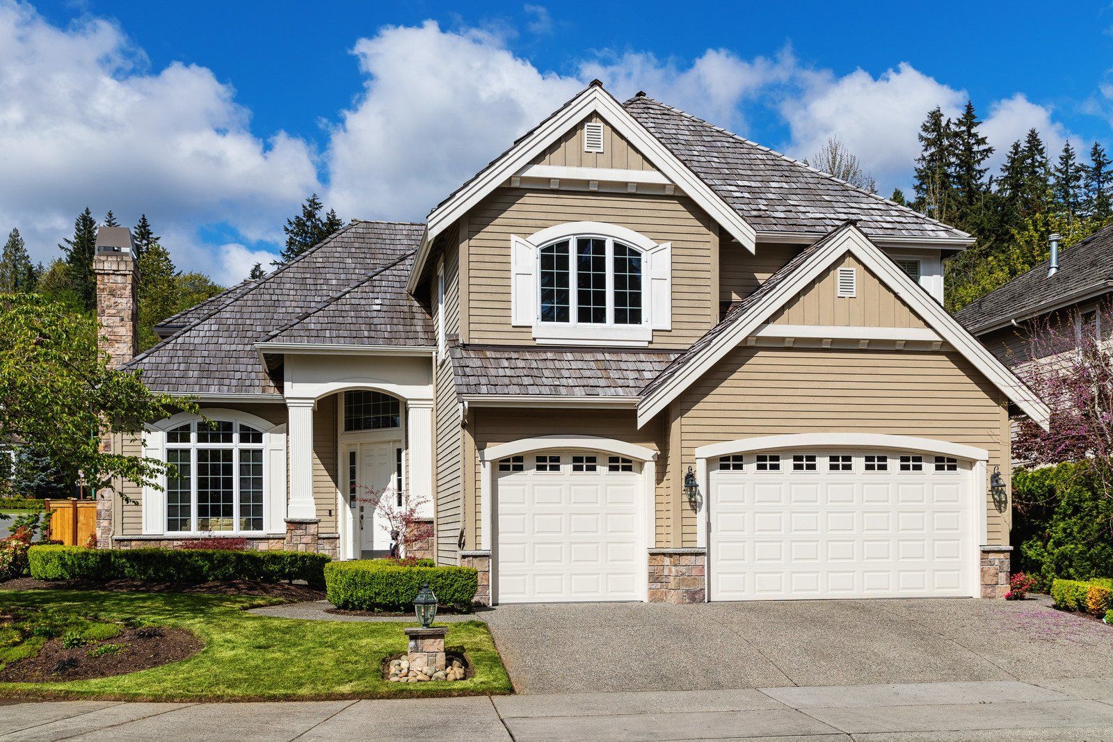 Two-story house with tan siding, gray roof, white garage doors, and a green lawn under a blue sky.
