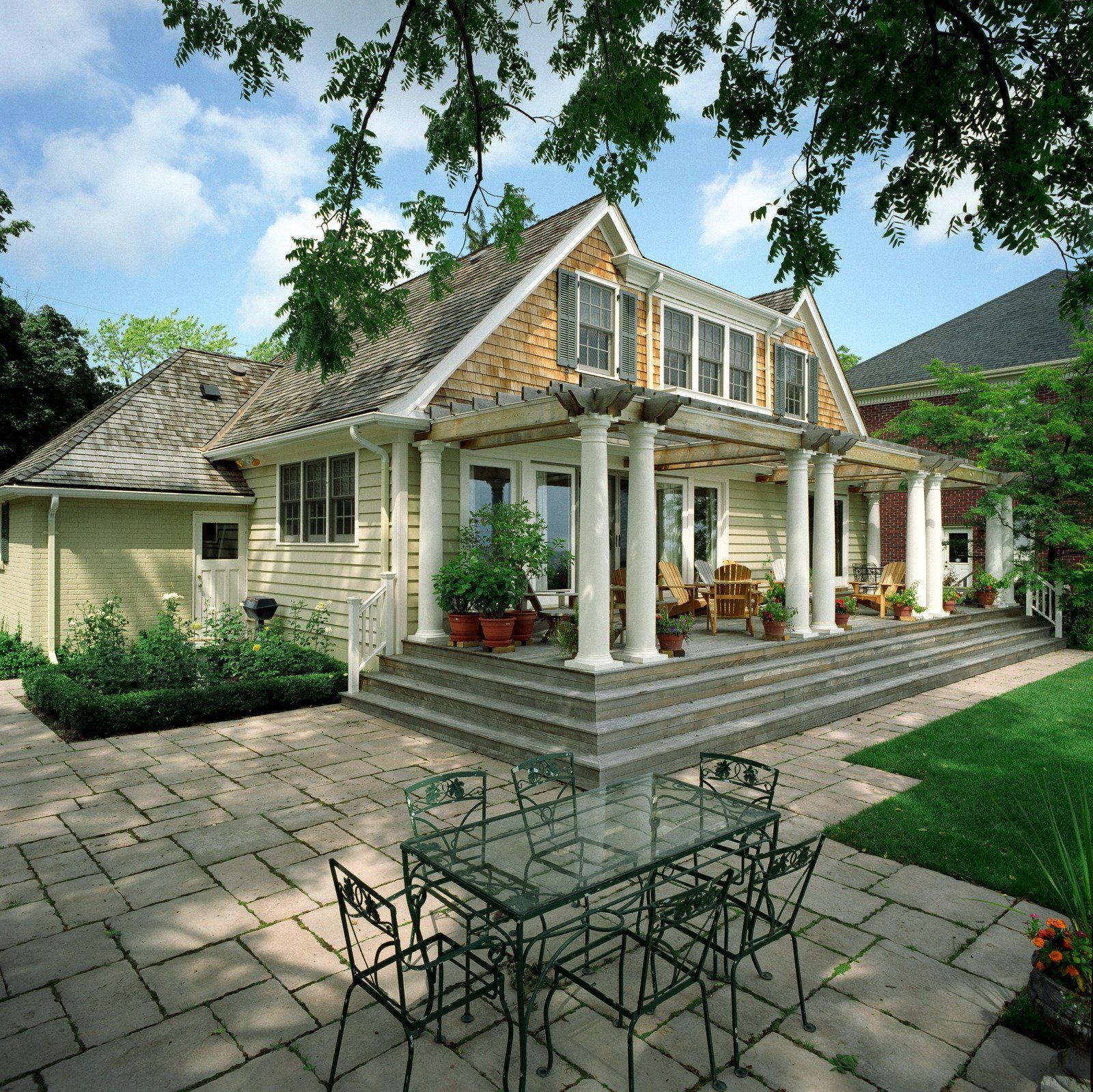 House with a stone patio and porch with white columns, green lawn, blue sky.